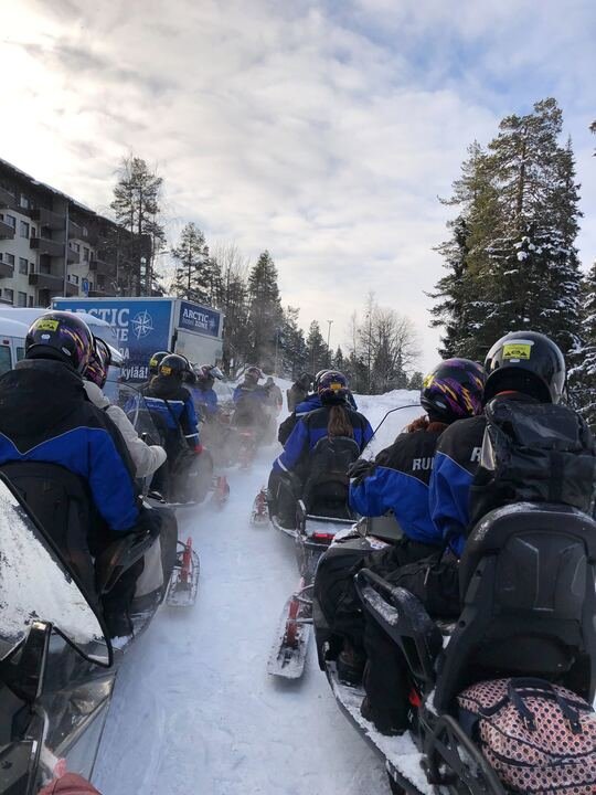 Group of people riding snowmobiles on a snowy trail surrounded by trees with cloudy sky overhead.
