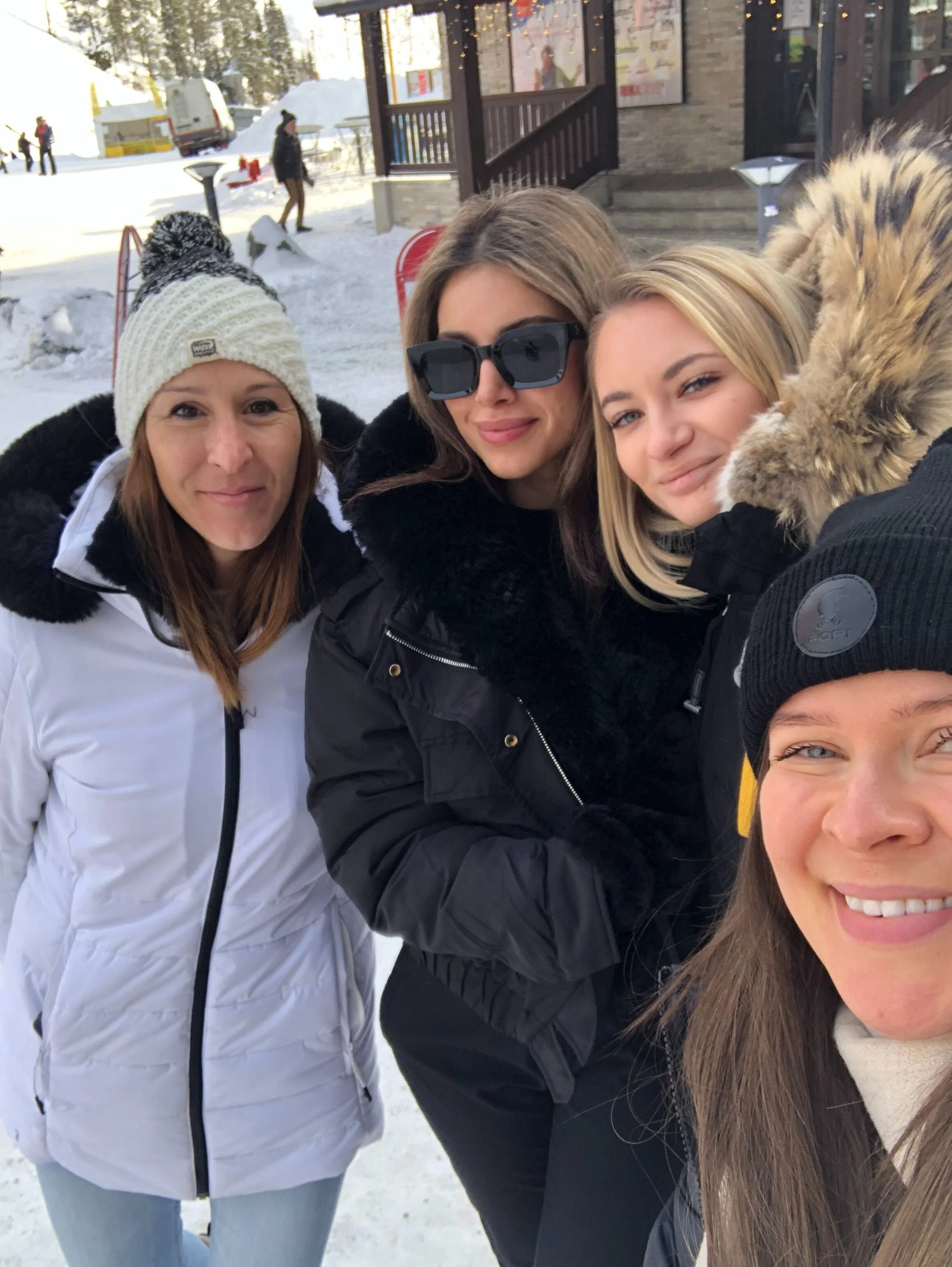 Four women in winter clothing taking a selfie outdoors in a snowy area with buildings and other people in the background.