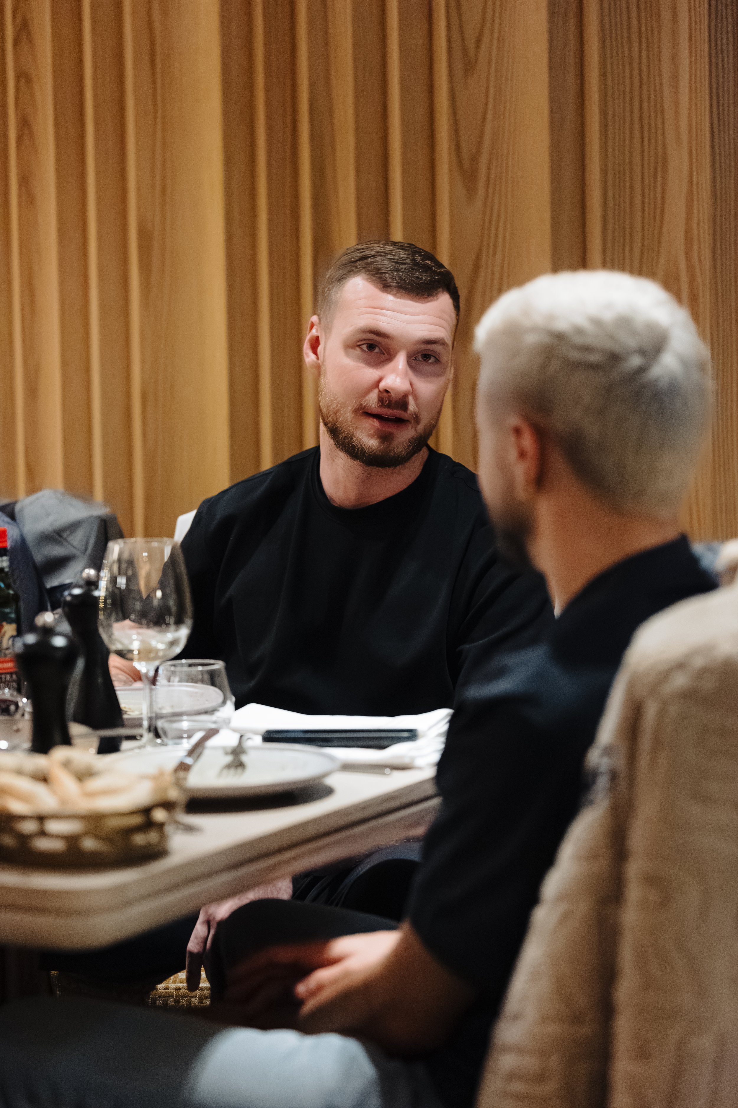 Two men having a conversation at a restaurant table. One man has short brown hair and a beard, wearing a black shirt, the other has platinum blond hair, wearing a black shirt and beige jacket. The table has plates, glasses, and condiments.