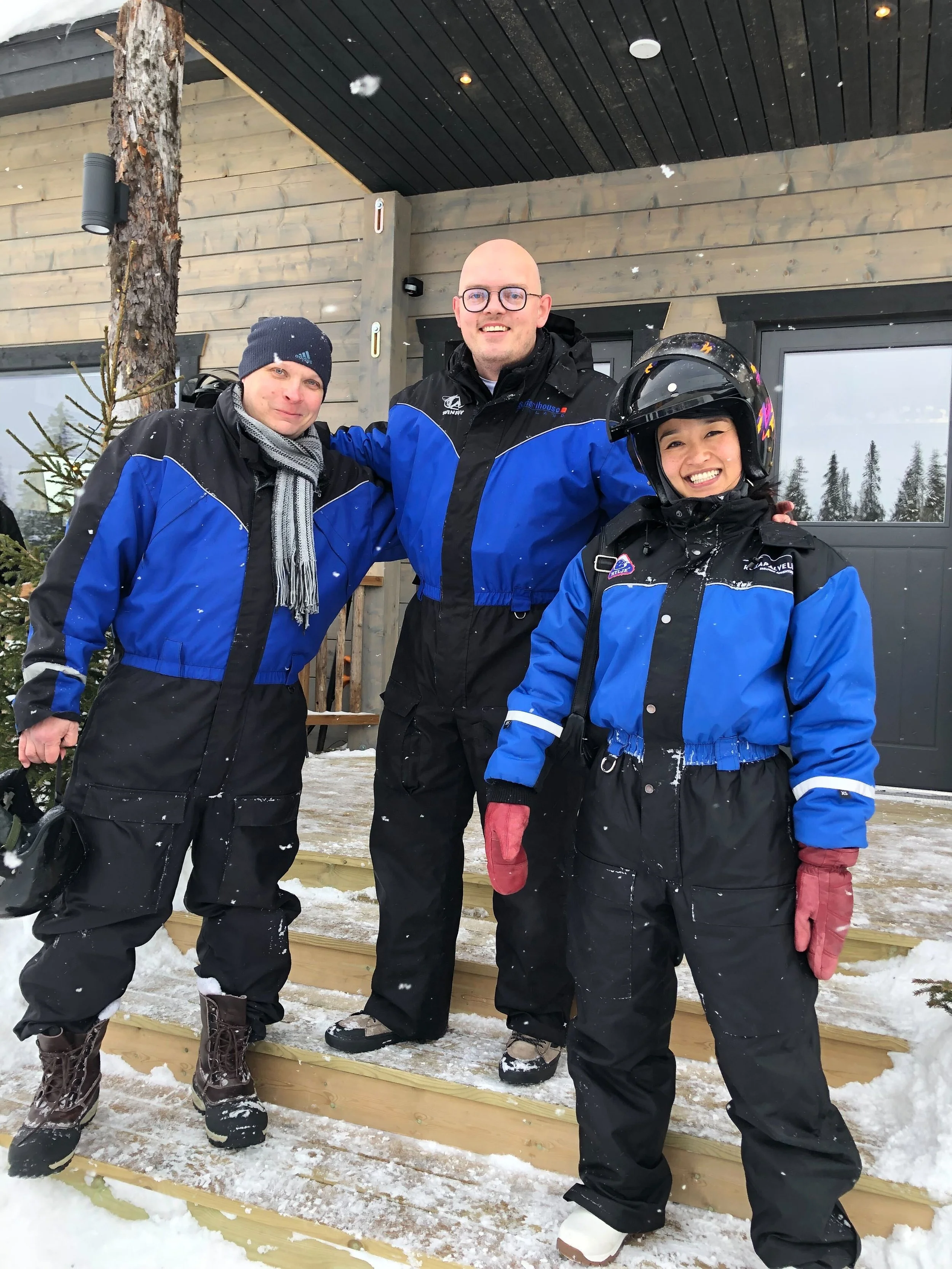 Three people in winter gear standing on snow-covered steps in front of a building, smiling at the camera, with snowy trees and a house in the background.