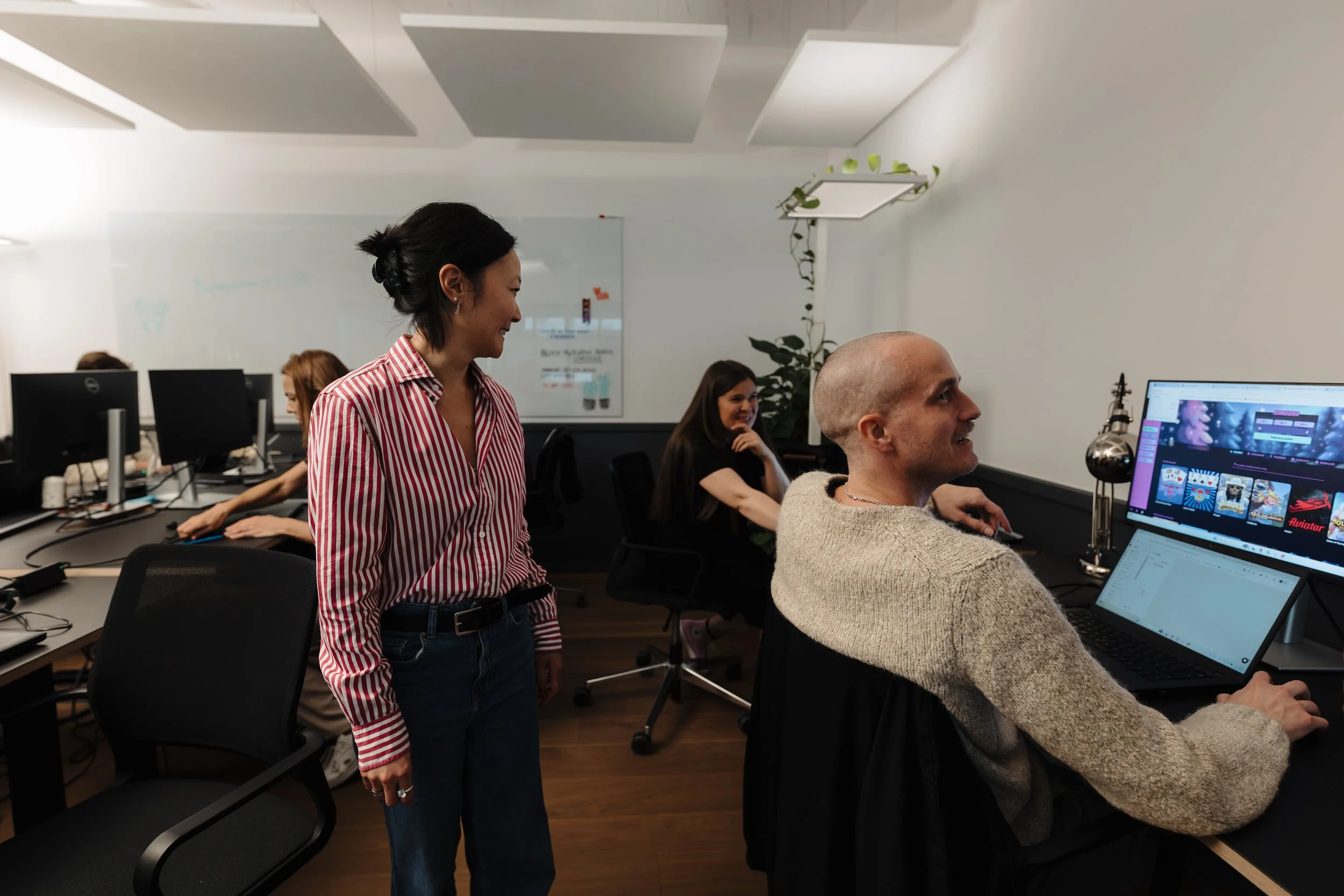 Office environment with four people working at their desks; two women and two men, one woman standing and smiling at a seated man.