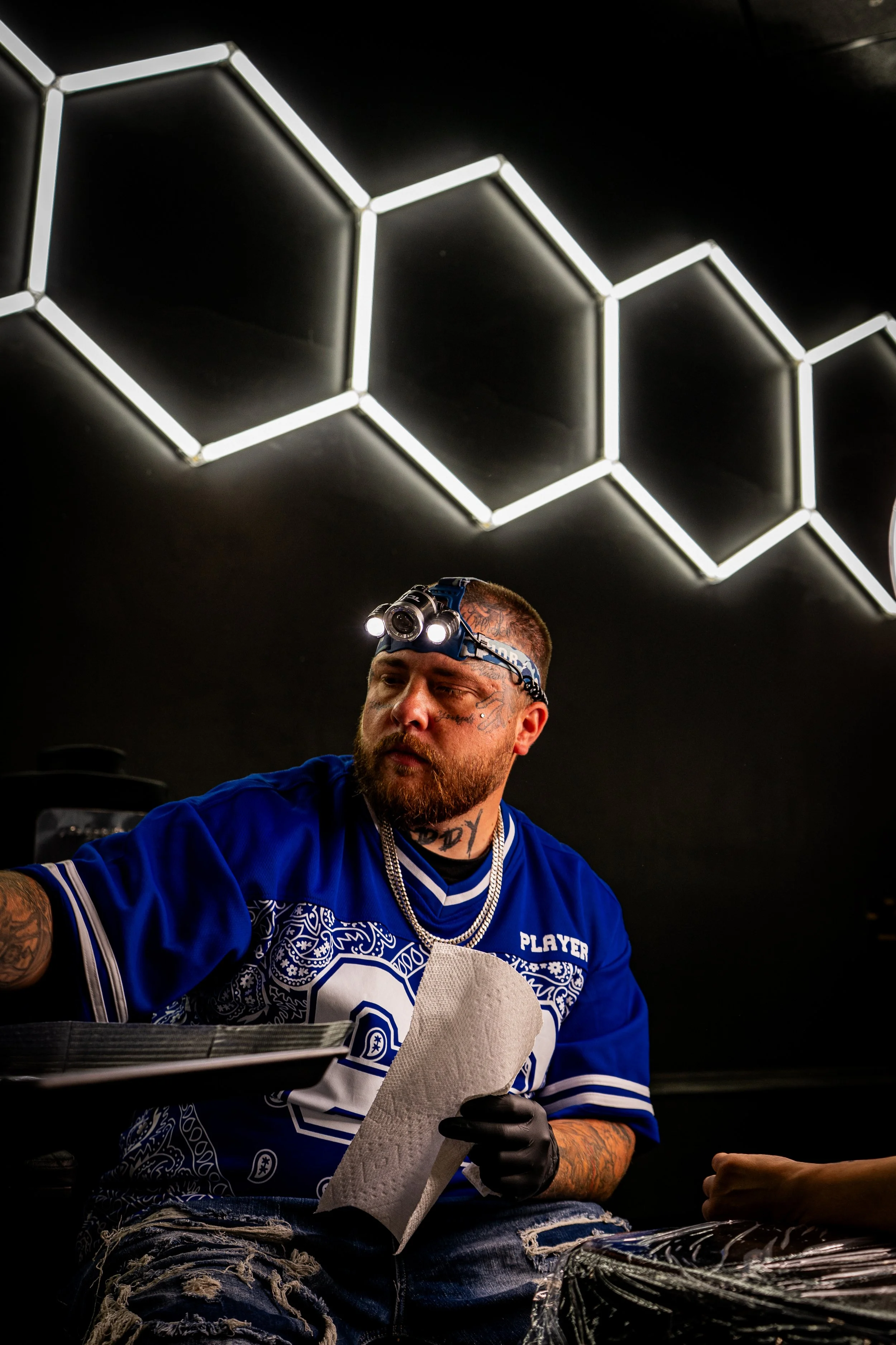 A man with tattoos and a beard wearing a blue sports jersey, black gloves, and a headlamp, sitting and handling a roll of paper towel in a dark room with a hexagonal neon light ceiling design.