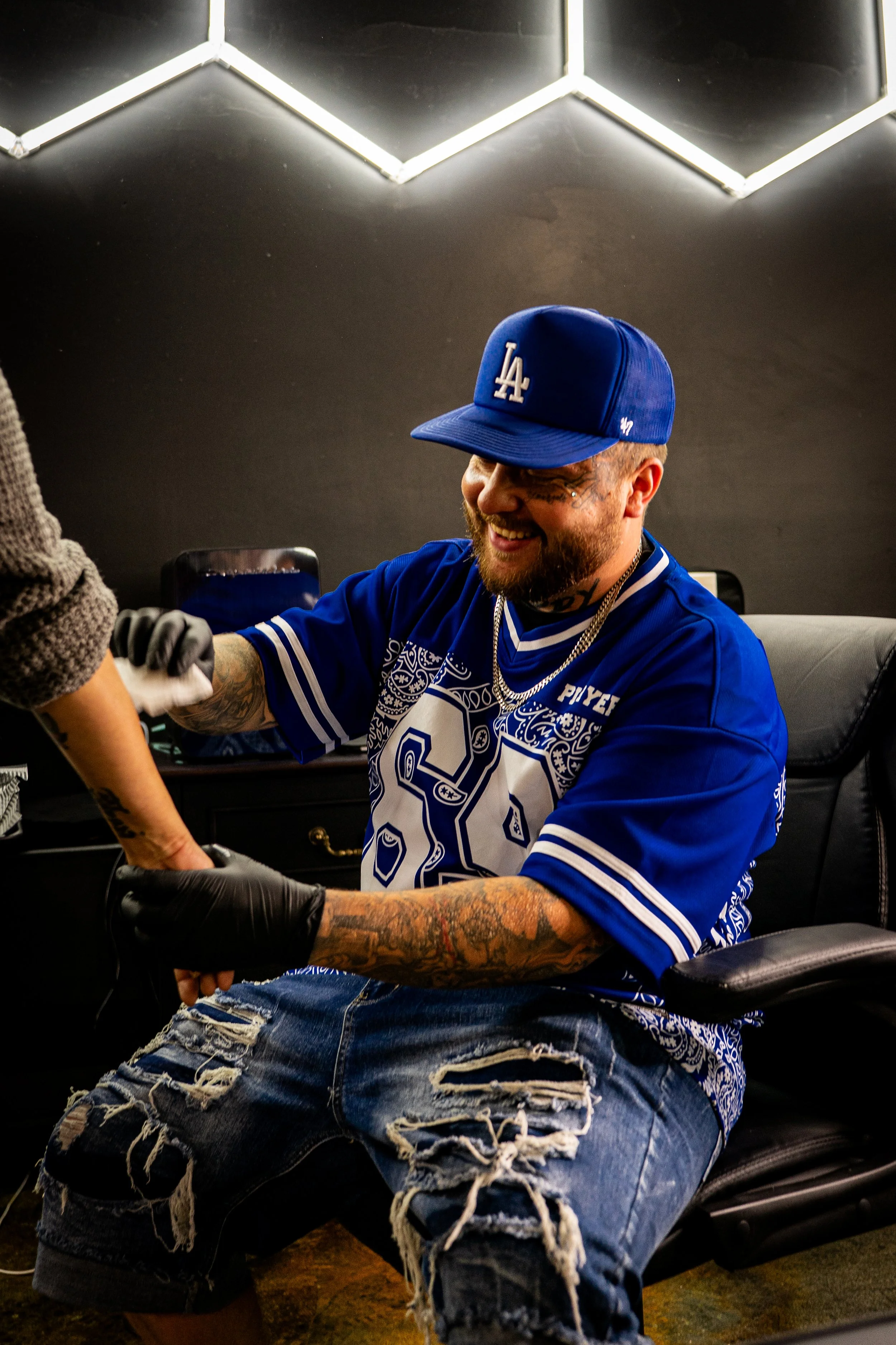 A man wearing a Los Angeles Dodgers cap, a blue sports jersey, and ripped jeans is smiling and shaking hands with someone wearing black gloves in an indoor setting with a black wall and hexagonal light fixtures overhead.