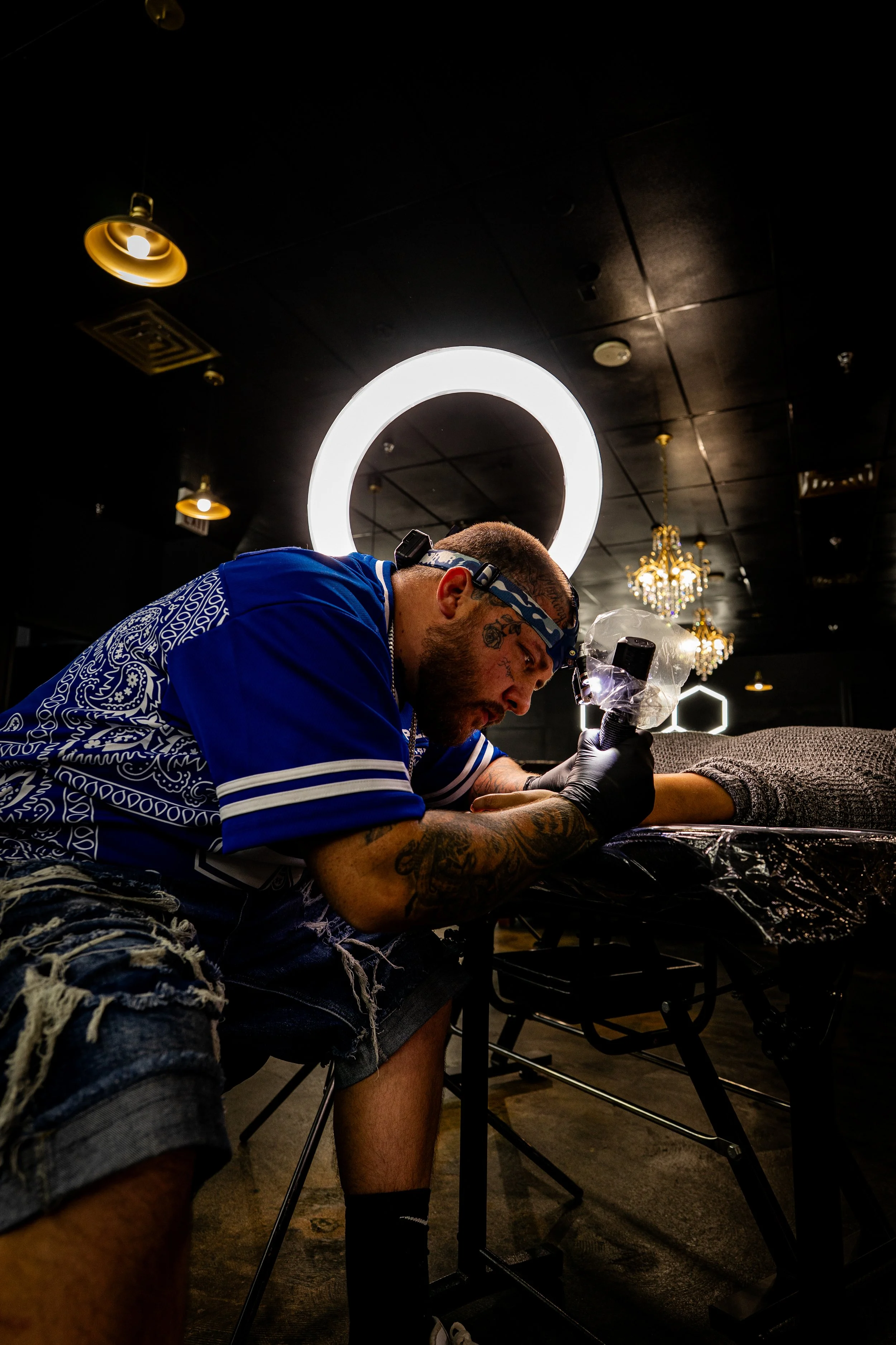 Tattoo artist tattooing a person's arm in a tattoo studio with modern lighting and chandeliers.