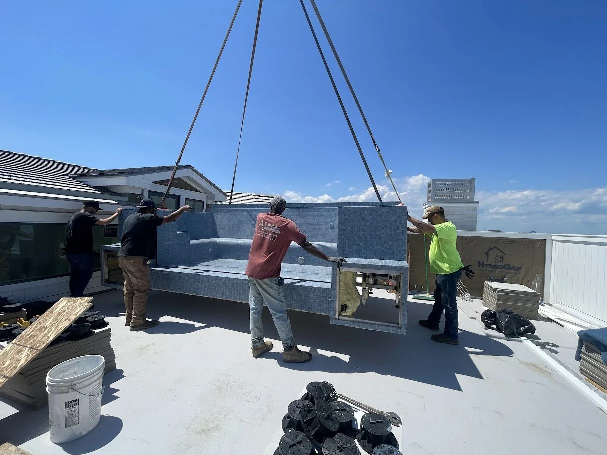 Four workers install a large, above-ground swimming pool on a rooftop under a clear blue sky.