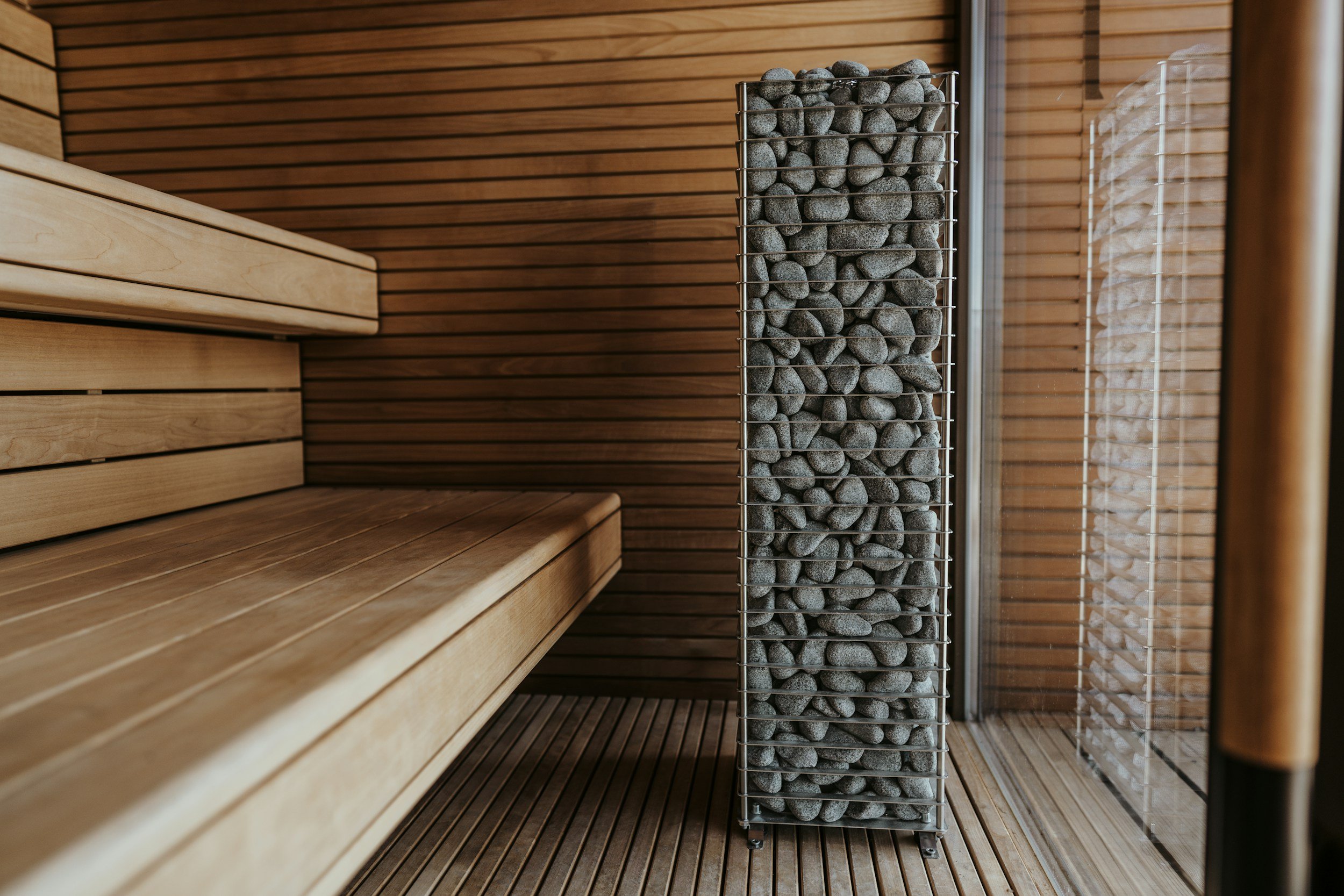 Interior of a sauna with wooden benches and a tall metal container filled with sauna stones.