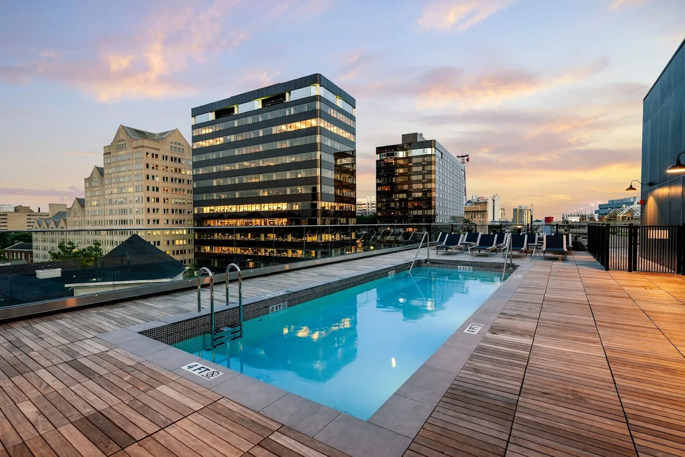 Rooftop swimming pool with lounge chairs overlooking city skyscrapers at sunset.