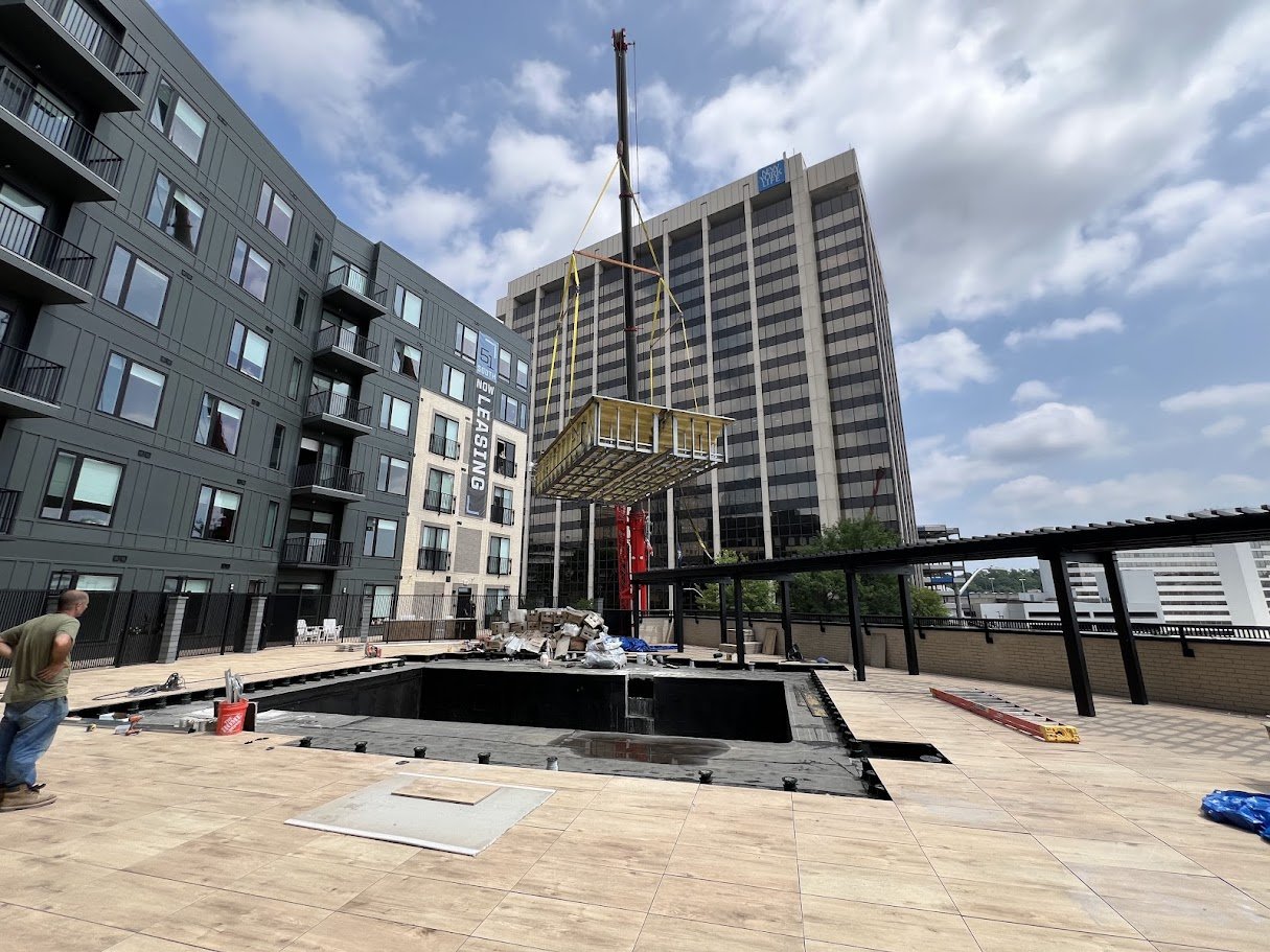 Urban construction site with a man observing, an open hole in the ground, construction materials, and a lift platform overhead, surrounded by modern apartment buildings and skyscrapers under a partly cloudy sky.