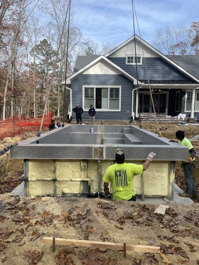 Construction workers installing a concrete foundation or pool in the backyard of a blue house with trees in the background.