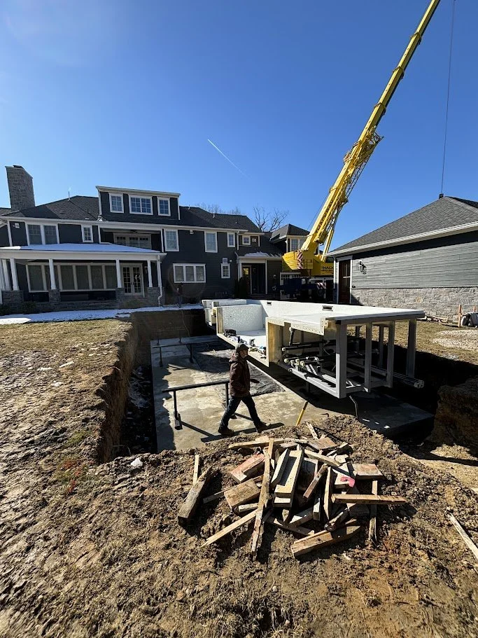 Construction site with a deep excavation for foundation, a worker walking across, and a large yellow crane lifting a prefabricated building element. Residential houses are visible in the background.