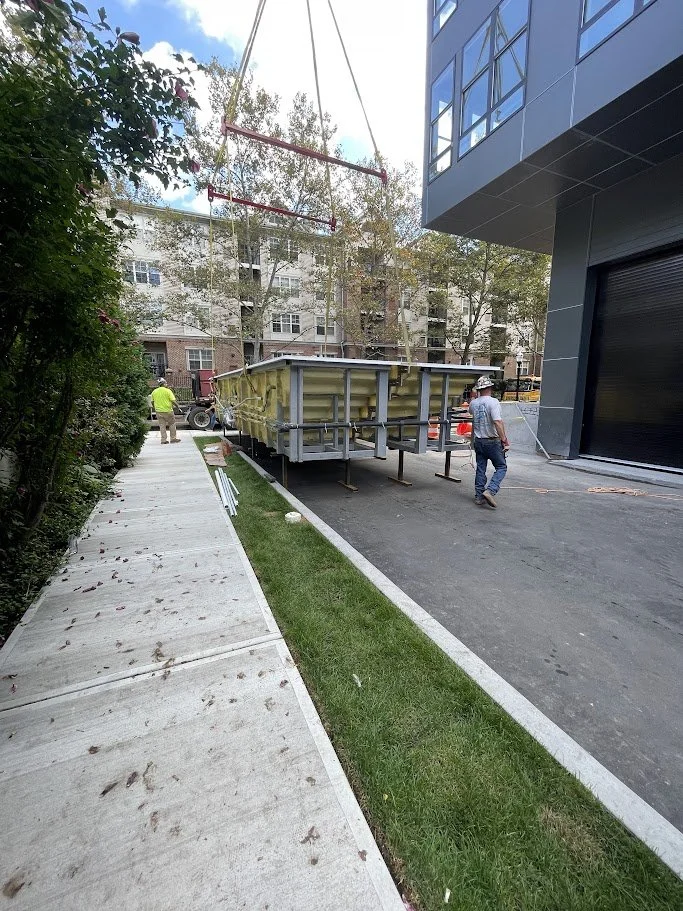 Construction workers are working on a new sidewalk and building exterior next to a modern commercial building, with a large construction trailer nearby and a swing set overhead.