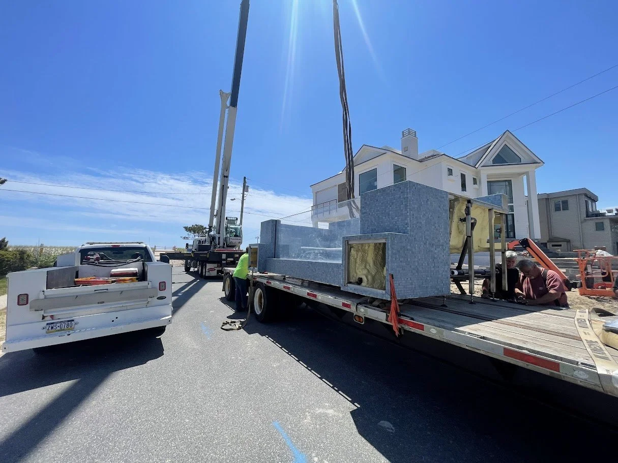 Construction workers are installing a large gray tiled hot tub or pool on a flatbed truck in front of a white multi-story house on a sunny day.