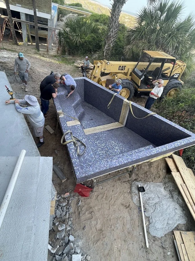 Workers installing a mosaic-tiled swimming pool at a construction site with palm trees in the background, a bulldozer, and construction tools.