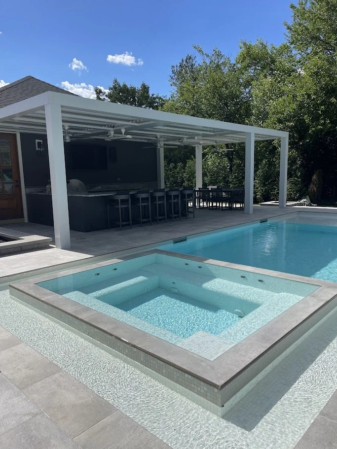 Backyard with swimming pool and hot tub, covered patio with outdoor kitchen and seating, surrounded by trees and blue sky.