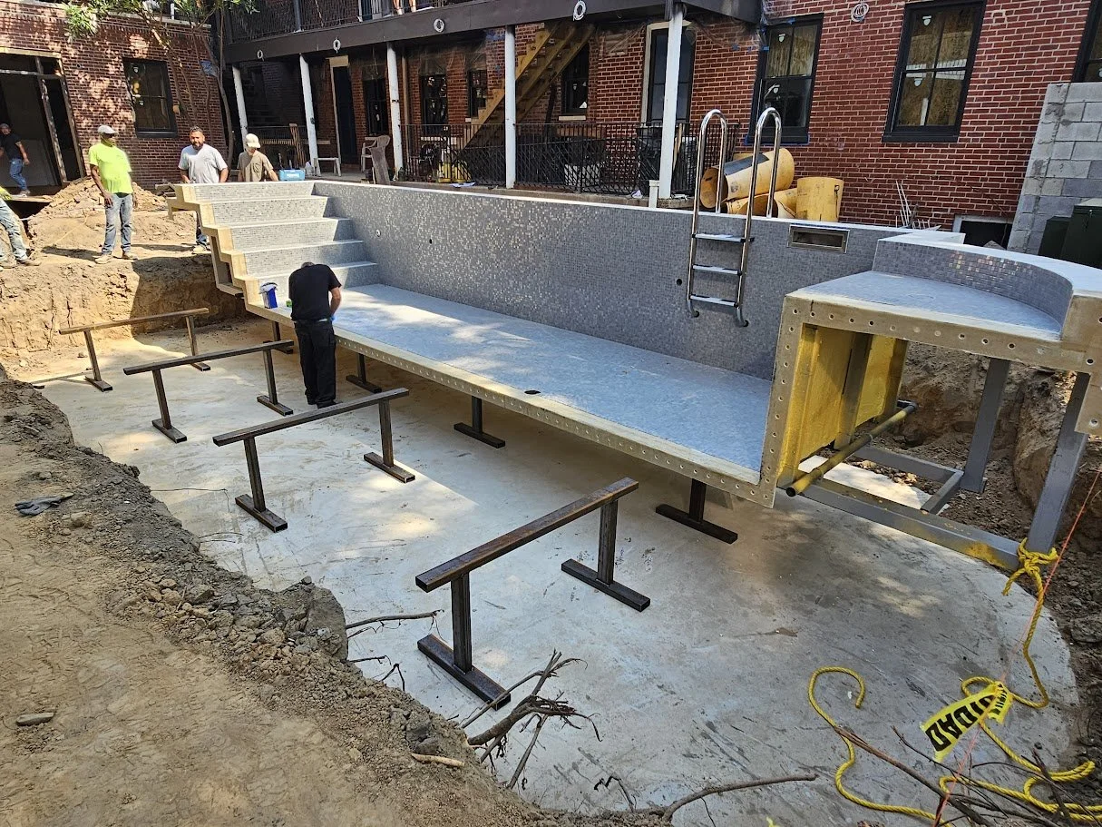 Construction workers building a swimming pool in a backyard, with a partially finished tile interior, a ladder, and surrounding safety barriers.