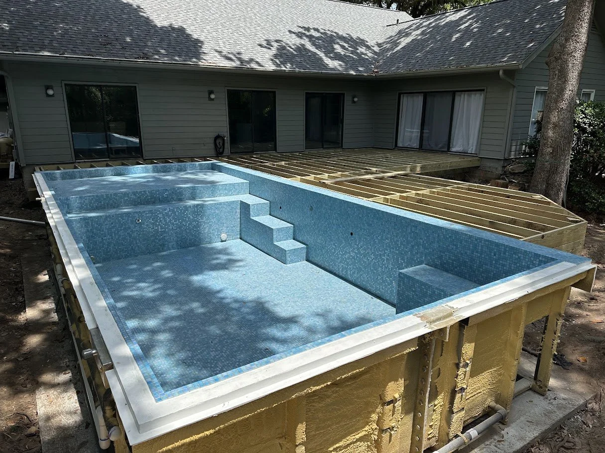 A residential backyard with an in-ground swimming pool under construction, with the pool's interior tiled in blue mosaic, next to an unfinished wooden deck, with a house in the background and trees casting shadows.