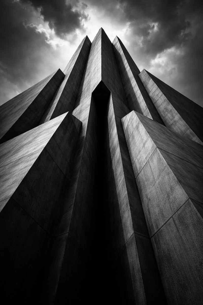 Black and white photo of a tall, modern building with sharp, angular lines and a dramatic sky with dark clouds overhead.