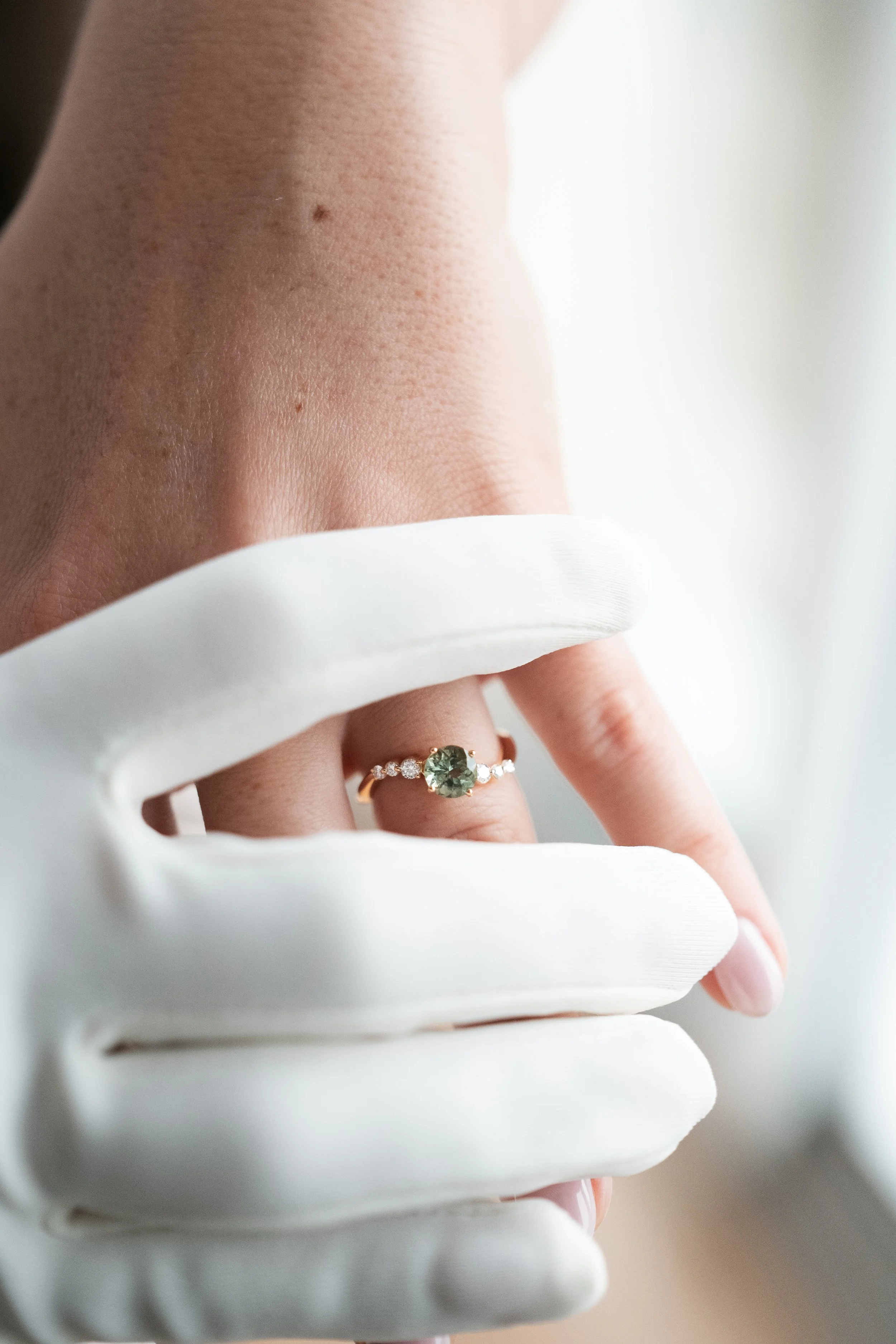 Main d'une personne portant une bague avec une pierre centrale verte et des petits diamants, portant aussi un gant blanc.