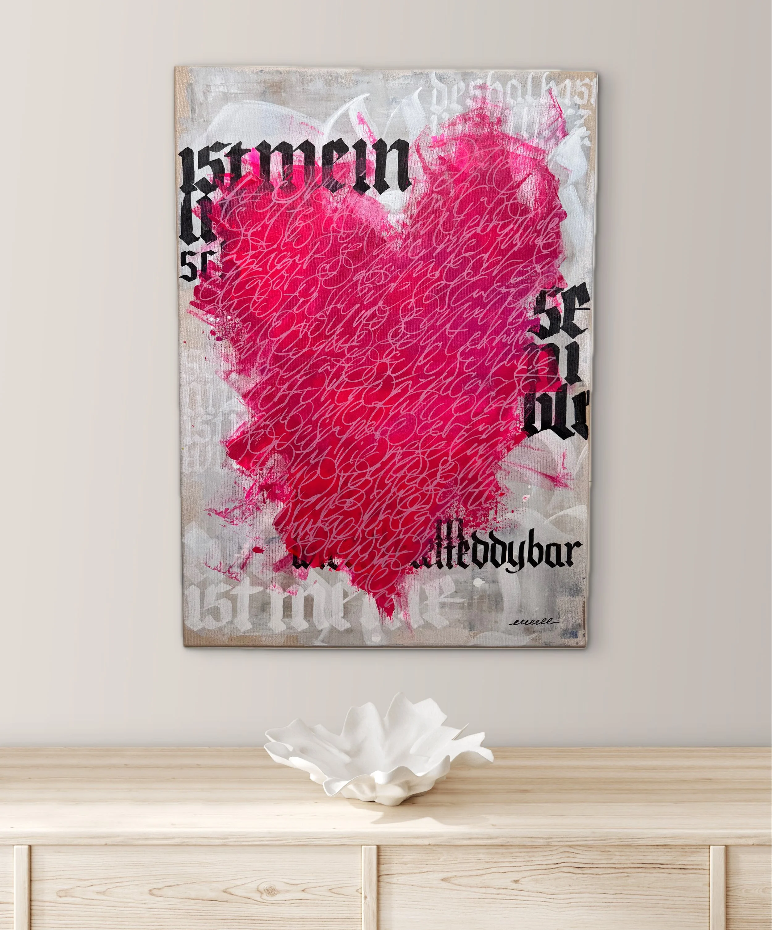 Cuadro con arte urbano que muestra un corazón rosa con escritura en cursiva, texto en estilo gótico y fondo de colores neutros, colocado sobre una pared blanca con una pieza decorativa blanca en la mesa.