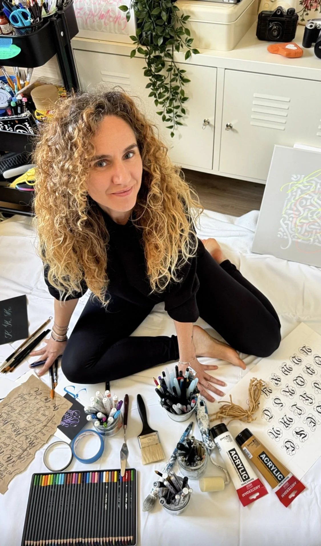 Mujer con cabello rizado y rubio sentado en una mesa con materiales de arte, rodeada de papeles, marcadores, pinceles y otros herramientas creativas, en un espacio que parece un estudio o una oficina.