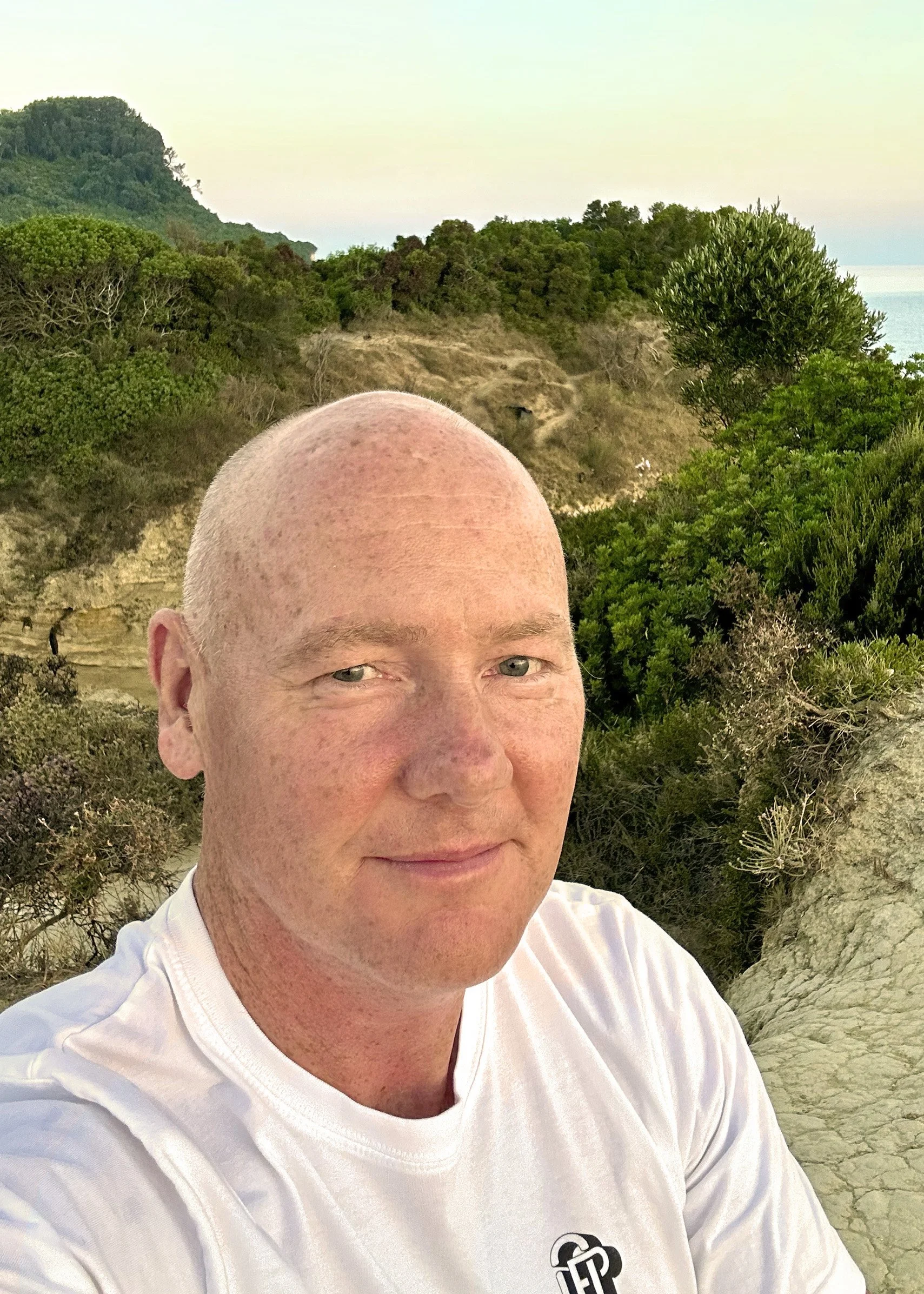 A man with a shaved head and freckles in a white t-shirt takes a selfie outdoors with a background of greenery, hills, and the ocean.