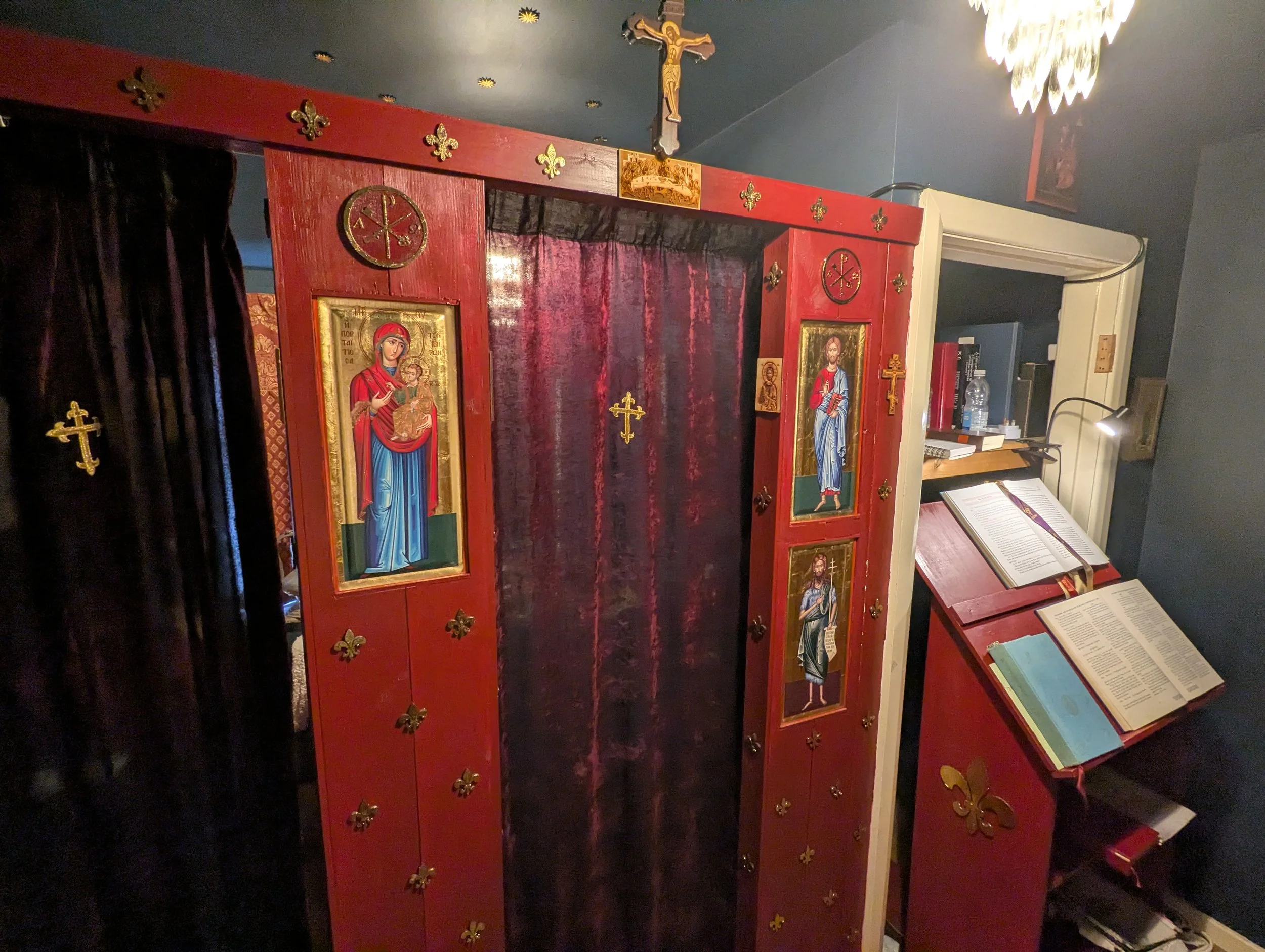 An ornate Catholic confession booth with red wood and gold accents, religious icons of Mary and Jesus, and a book stand with liturgical texts, inside a dimly lit church.