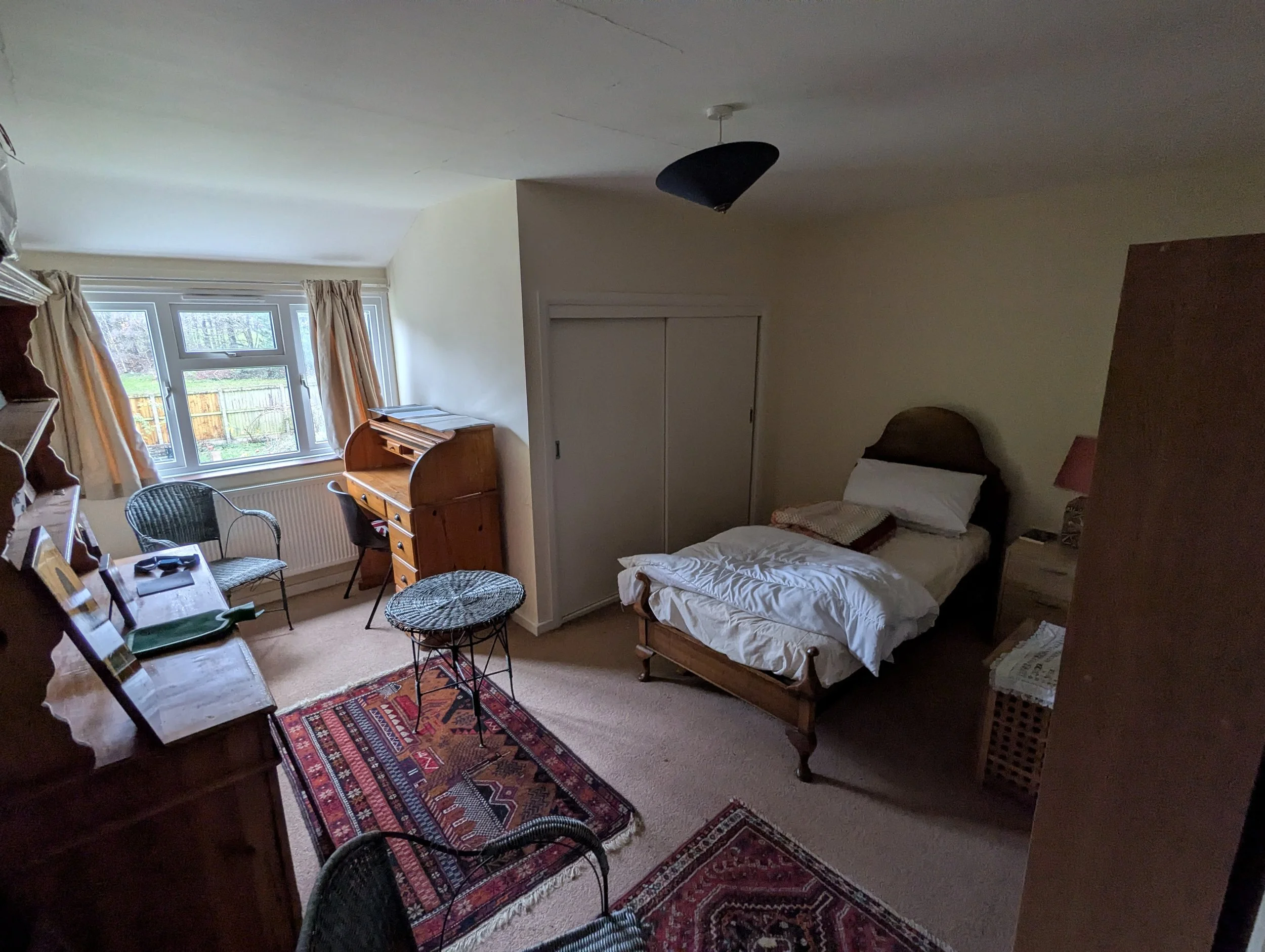 A cozy bedroom with a single bed, nightstand, and a small cabinet. There are chairs and a small table near the window, with a colorful rug on the carpeted floor. The window has curtains, and there is a ceiling light fixture.