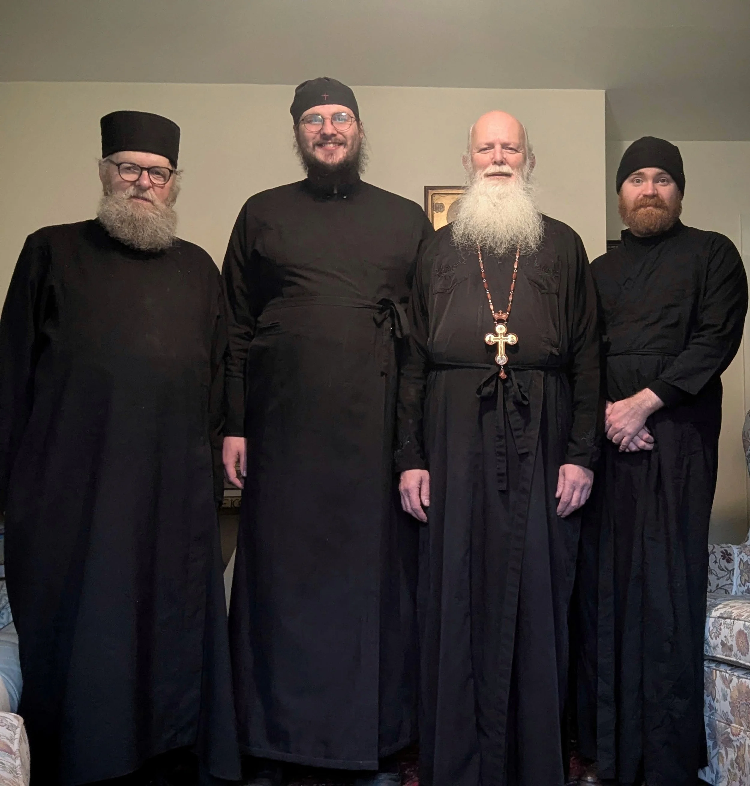 Four men dressed in black religious or clergy robes standing indoors, with two older men with long white beards and two younger men with short beards, all smiling.