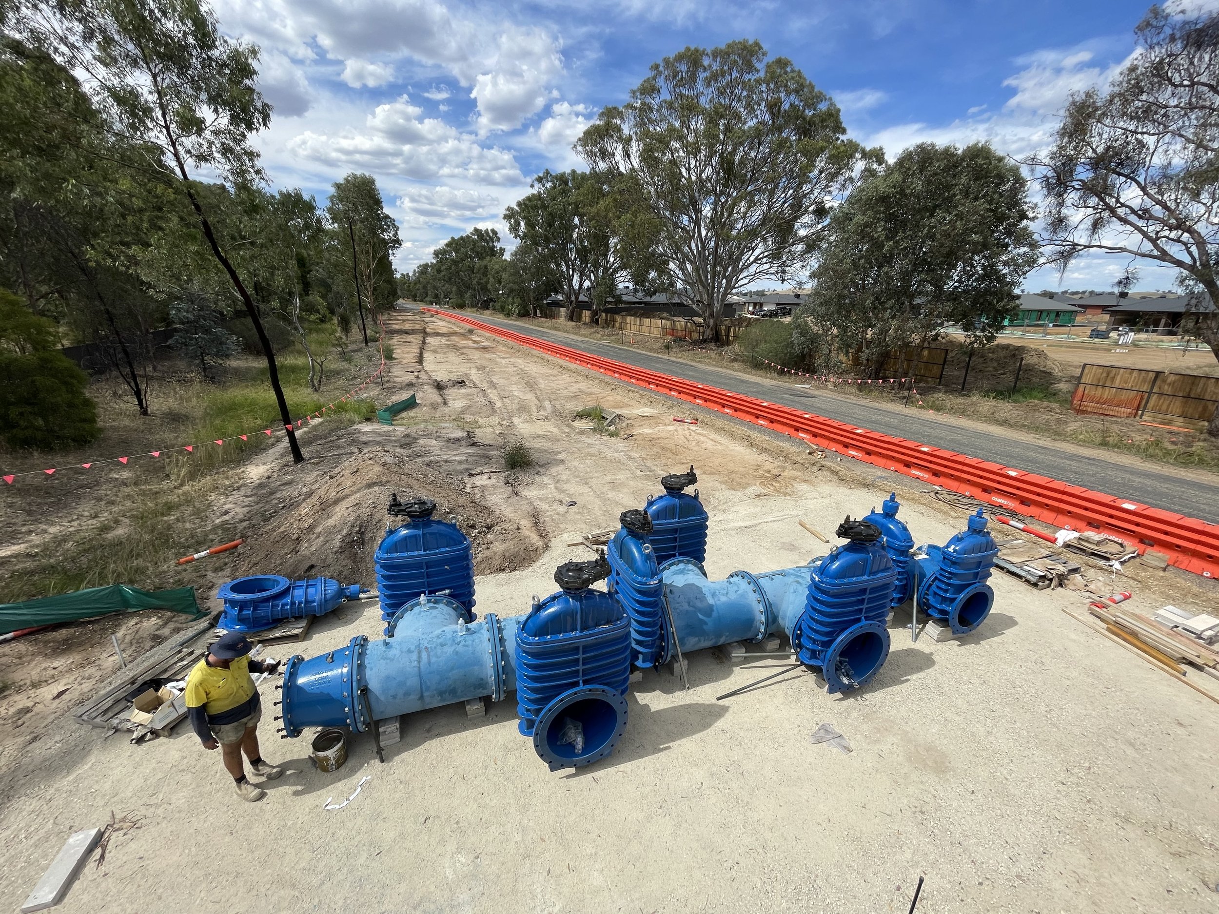 Construction site with large blue pipes and orange construction barriers along a dirt road with trees