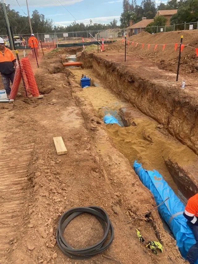 JEPC team install underground pipes in a trench at a construction site, with orange safety fencing and equipment nearby.