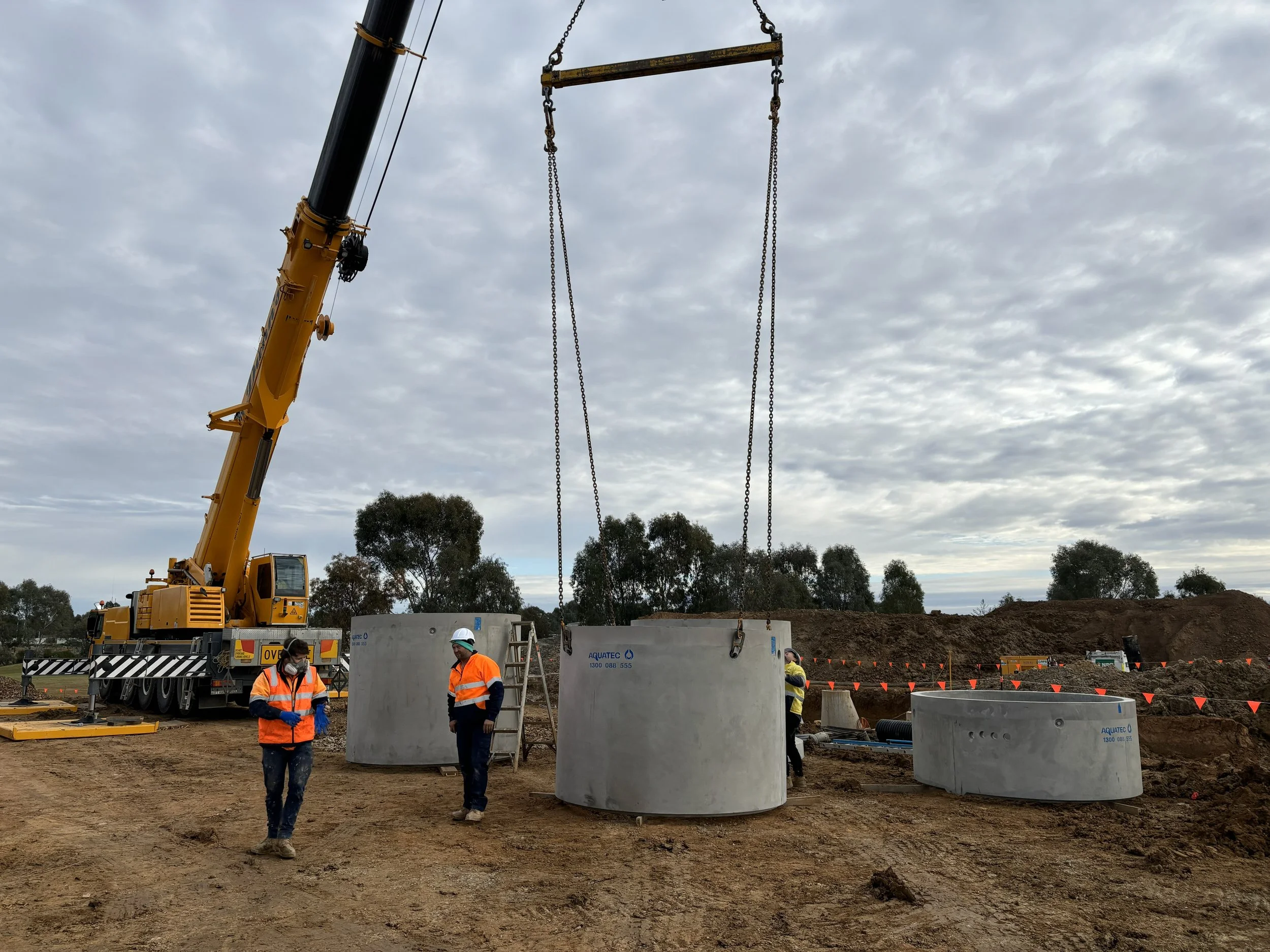 JEPC Construction site with a large yellow crane lifting concrete underground utility vaults while workers in orange safety vests and helmets observe and assist.