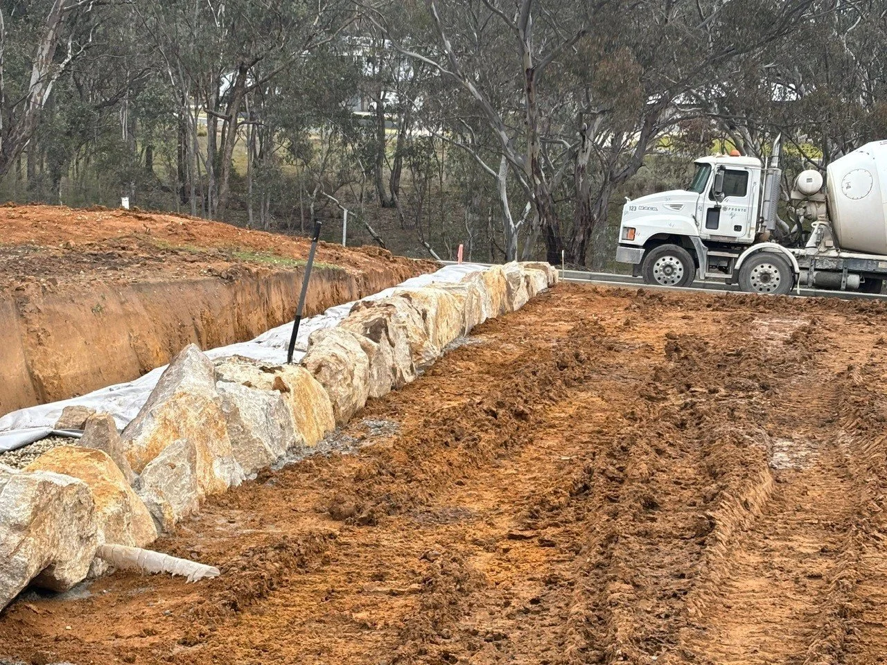 Construction site with a row of large rocks placed along a trench, and a cement mixer truck in the background