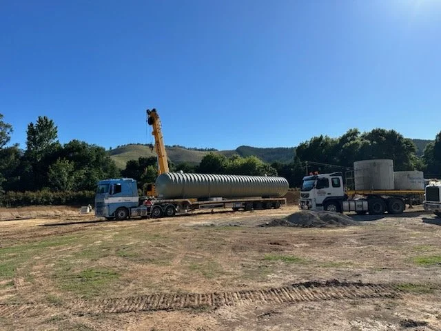 Construction site with two JEPC trucks and a crane lifting a large cylindrical tank 
