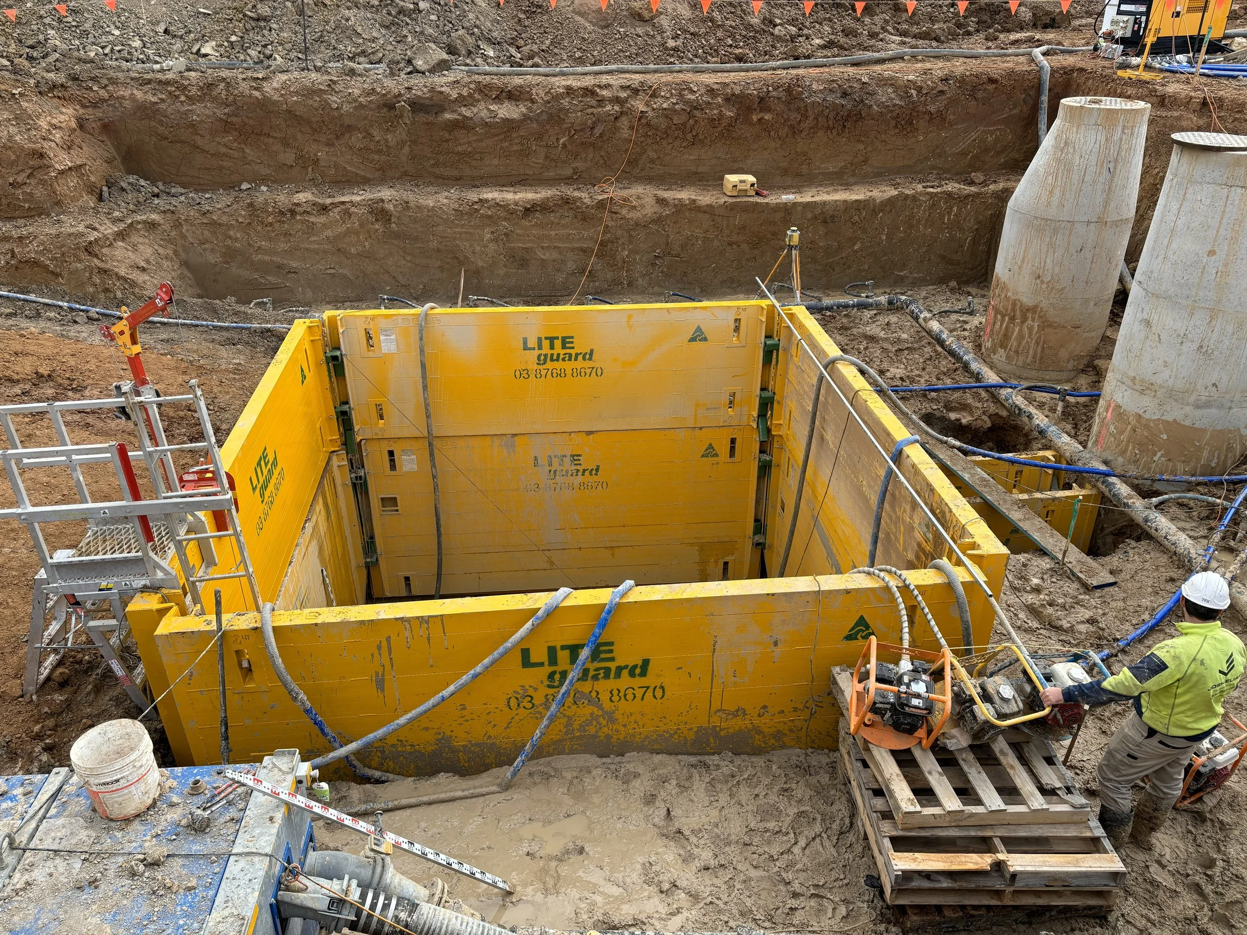 Construction site with excavation, yellow formwork labeled 'LITE guard', two large concrete pipes, a worker wearing a safety helmet and vest operating machinery, and various pipes and equipment.