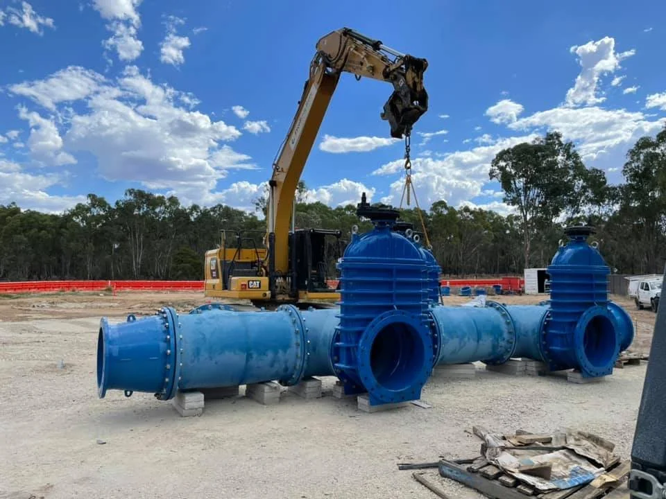 A large yellow excavator lifting a metallic blue valve for water infrastructure