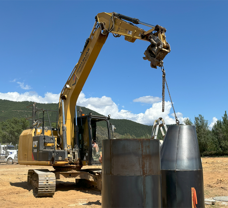 A yellow Caterpillar excavator lifting a large metallic cone-shaped object with a chain at a construction site.