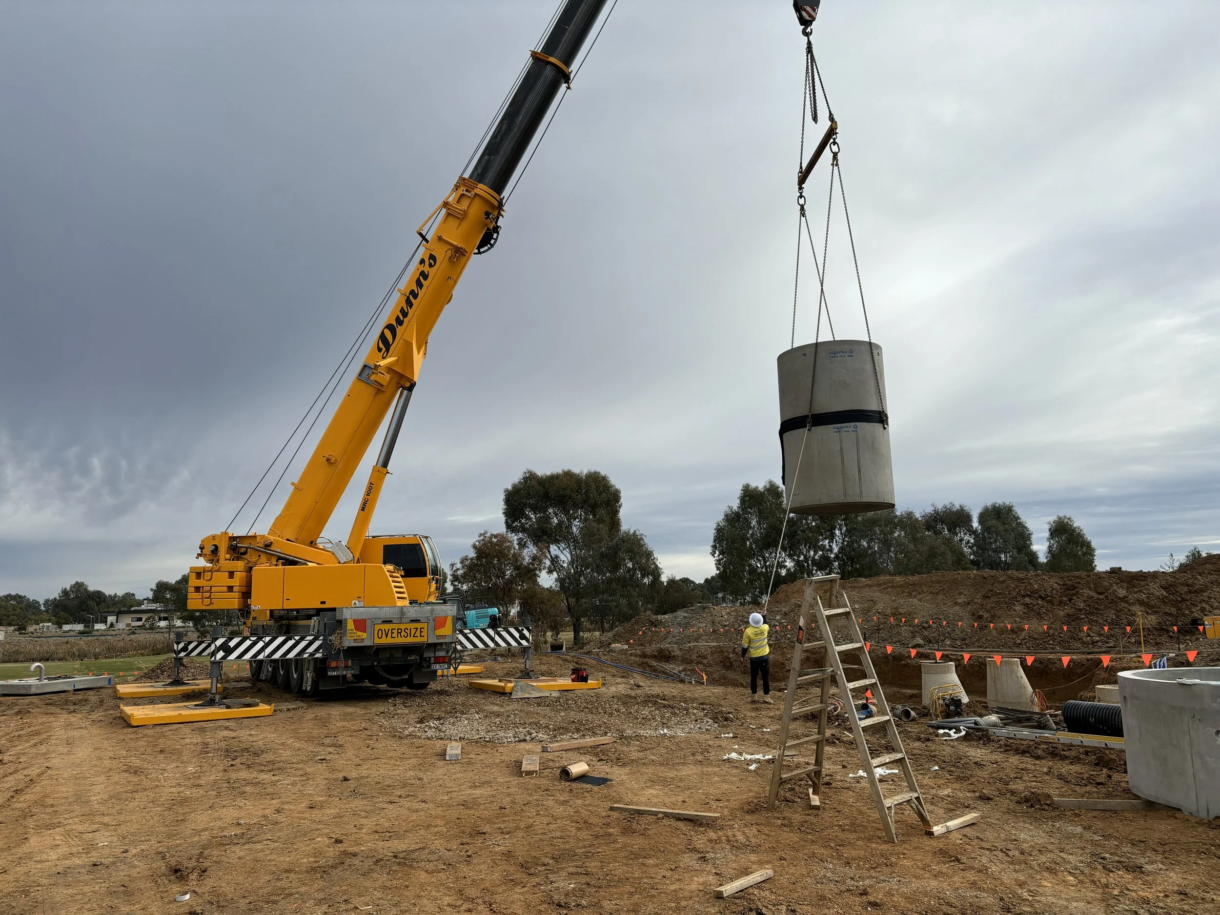 A construction site with a large yellow crane lifting a concrete cylinder, with a worker standing nearby