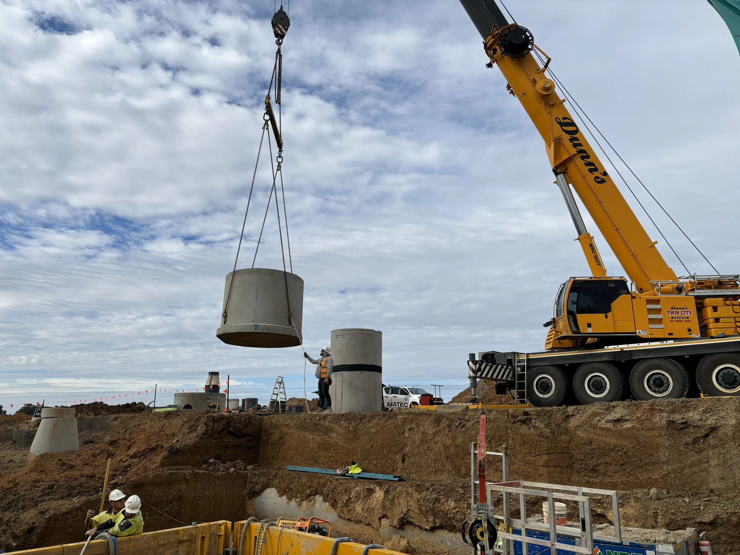 JEPC construction workers guiding large concrete sewer or drainage pipes being lifted by a yellow crane on an excavation site.