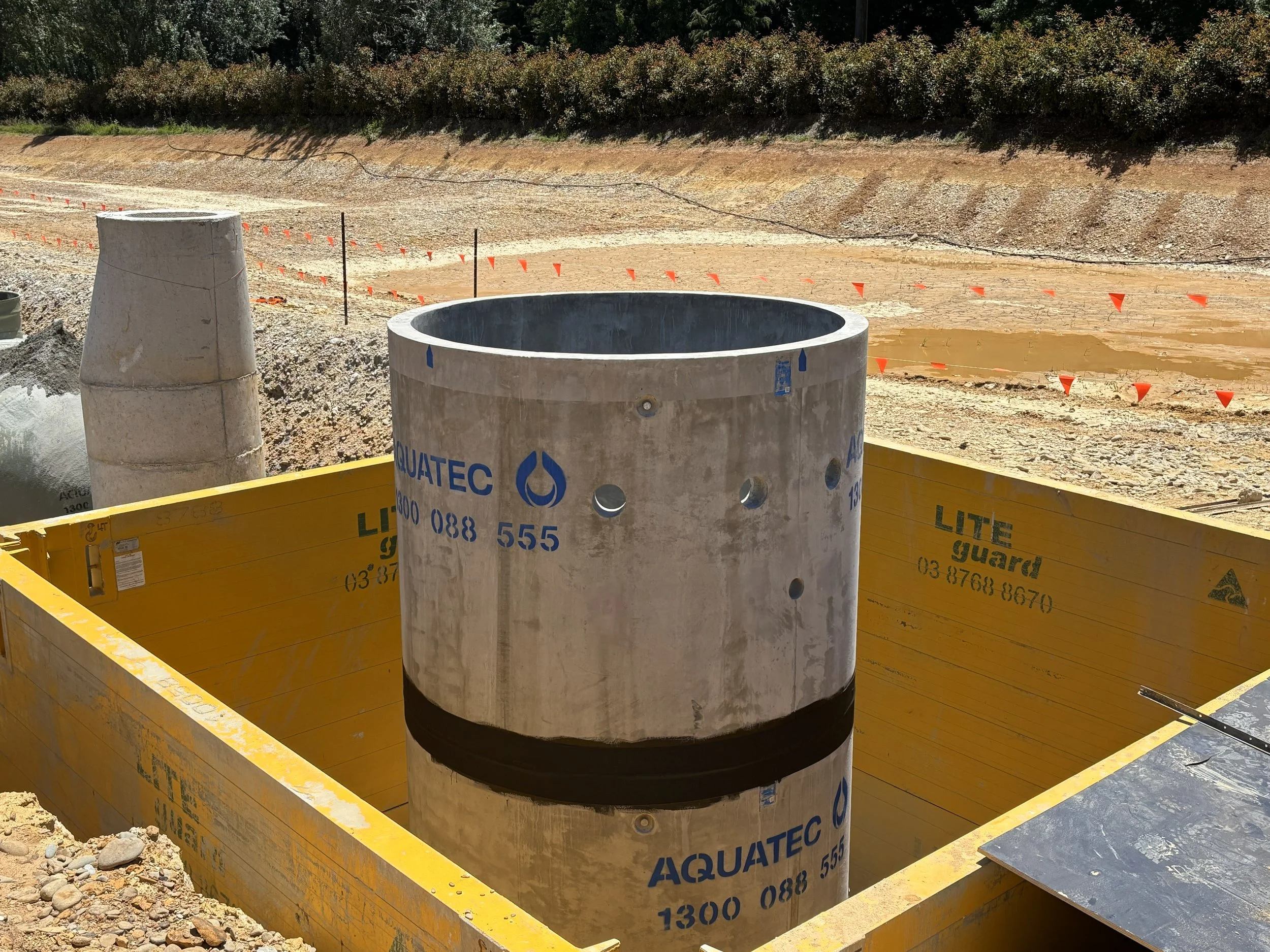 A cylindrical concrete pipe in a construction site, placed above a yellow waterproof barrier wall, with another pipe and construction materials visible in the background.
