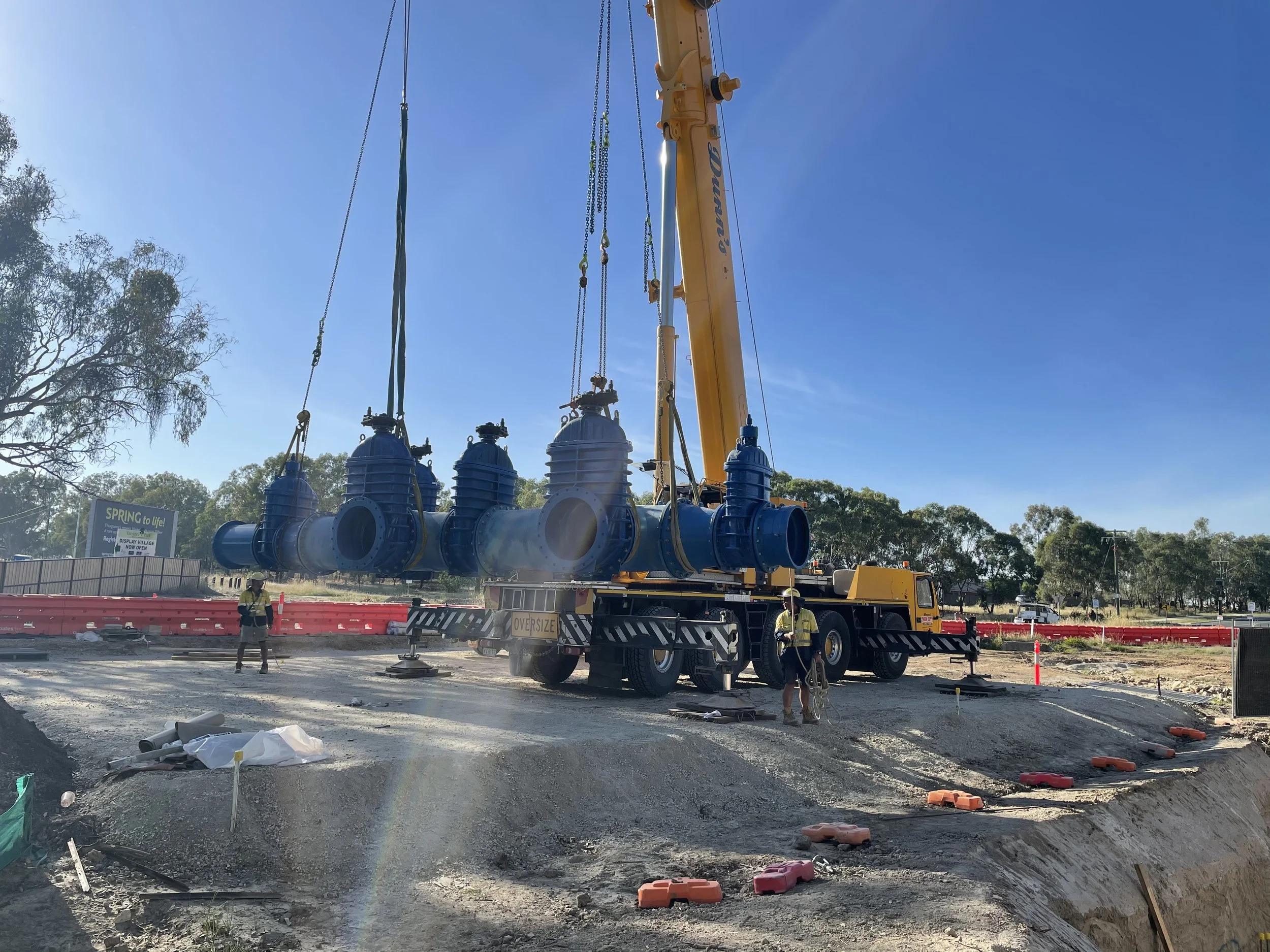 Construction workers operating a crane lifting large blue industrial pipes at a construction site on a sunny day.