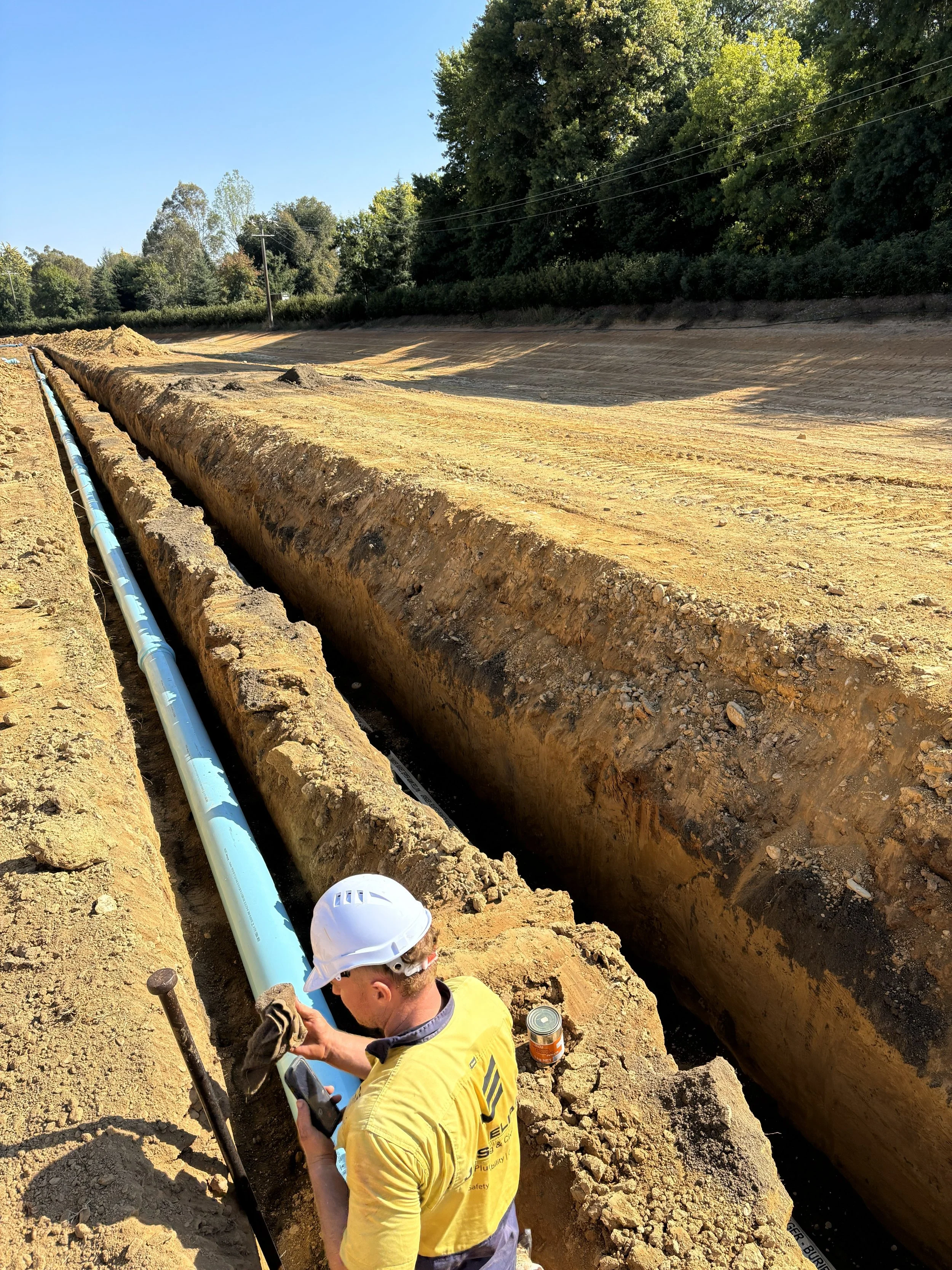 JEPC worker working in a trench with blue pipes installed, surrounded by excavated soil, with trees and power lines in the background