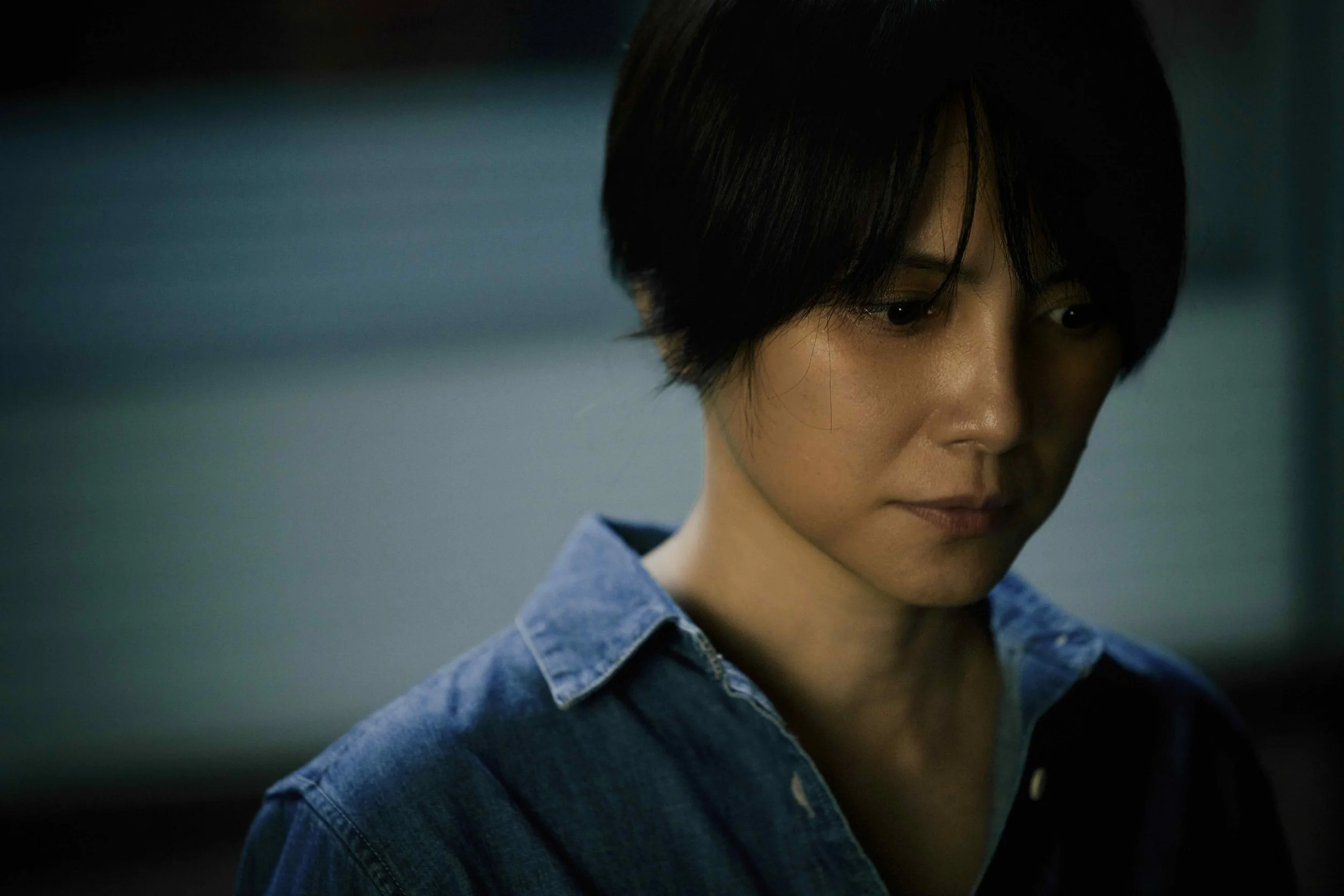 Close-up of a woman with short black hair looking down, wearing a denim shirt against a dark blurred background.