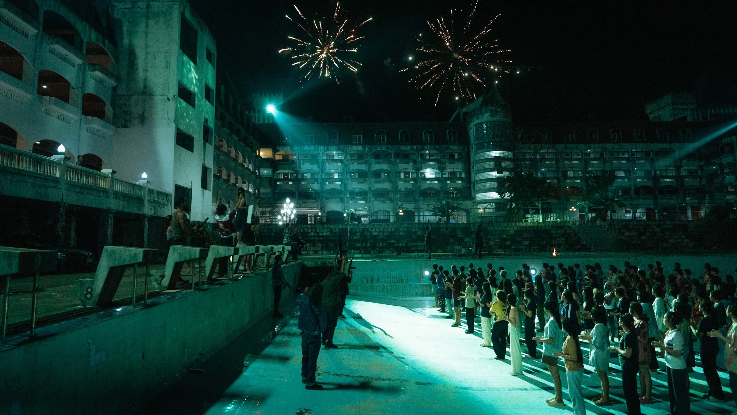 People gathered by an outdoor pool during fireworks celebration at night, with a large multi-story hotel or resort building in the background.