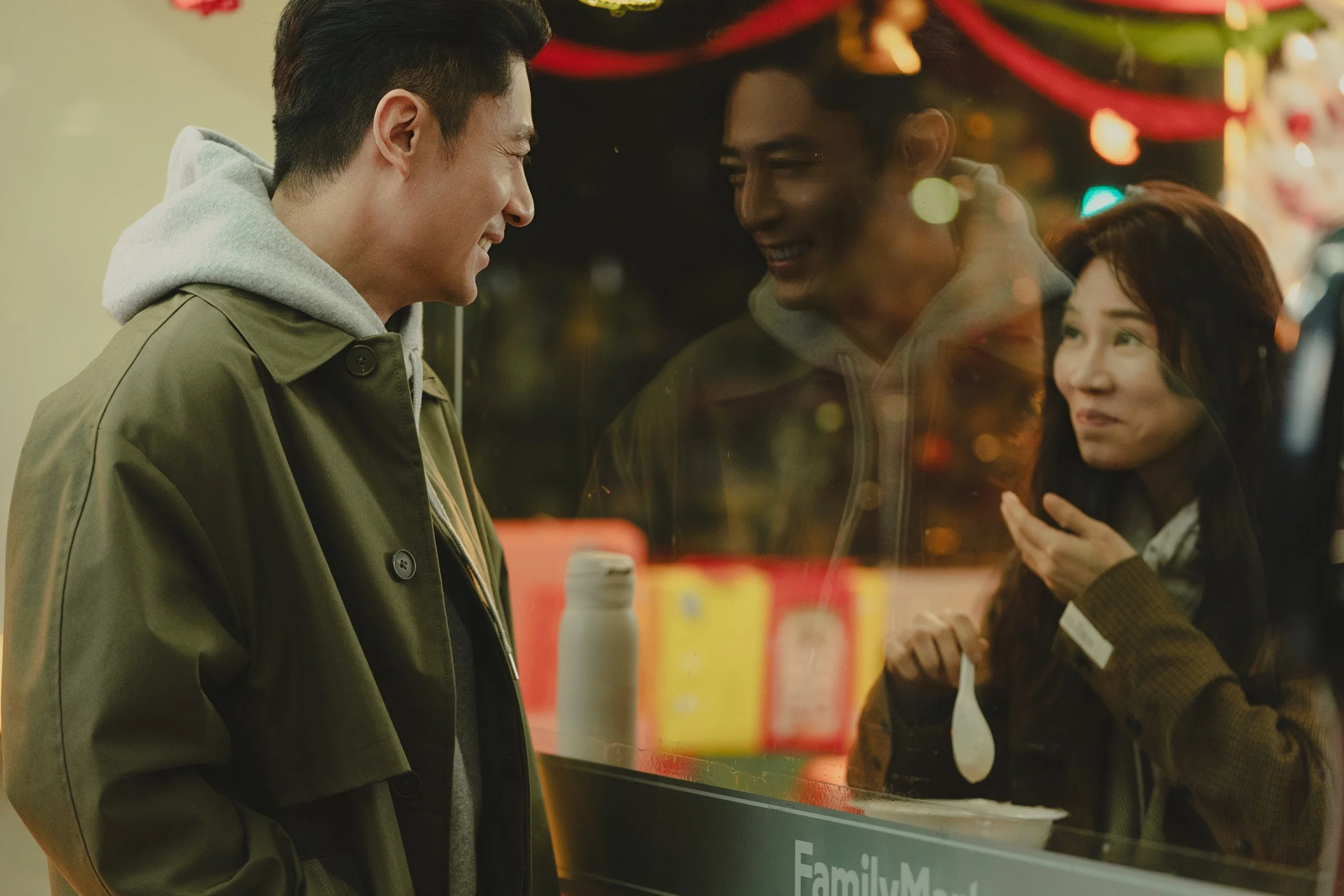 A young man and woman are talking through a glass window at a food stand, smiling and enjoying each other's company, with reflections of colorful lights in the glass.