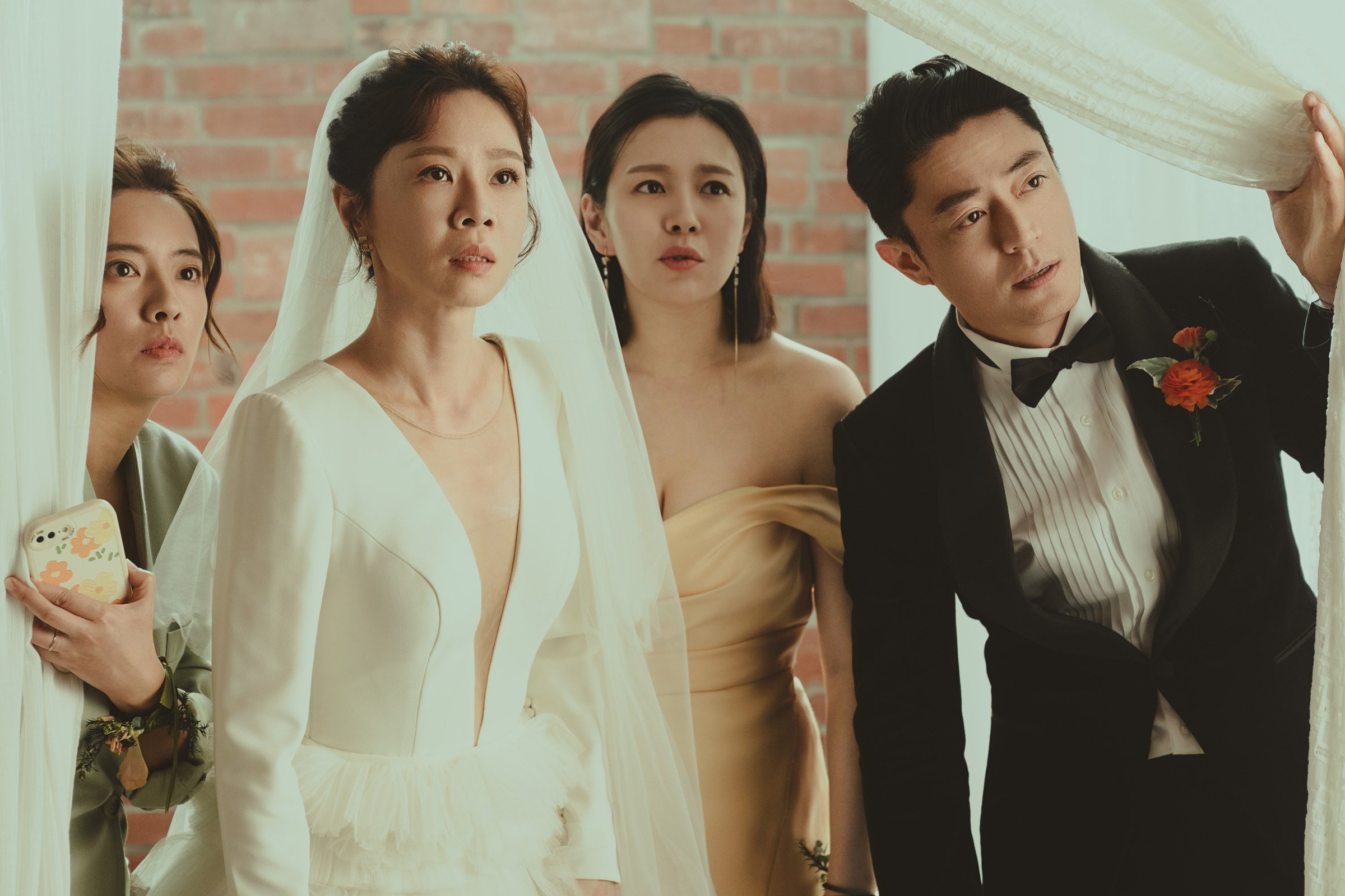 Four people peering through a curtain. Two women on the left in light-colored outfits, a woman in a beige strapless dress in the center, and a man in a black tuxedo on the right holding the curtain. The background features a brick wall.
