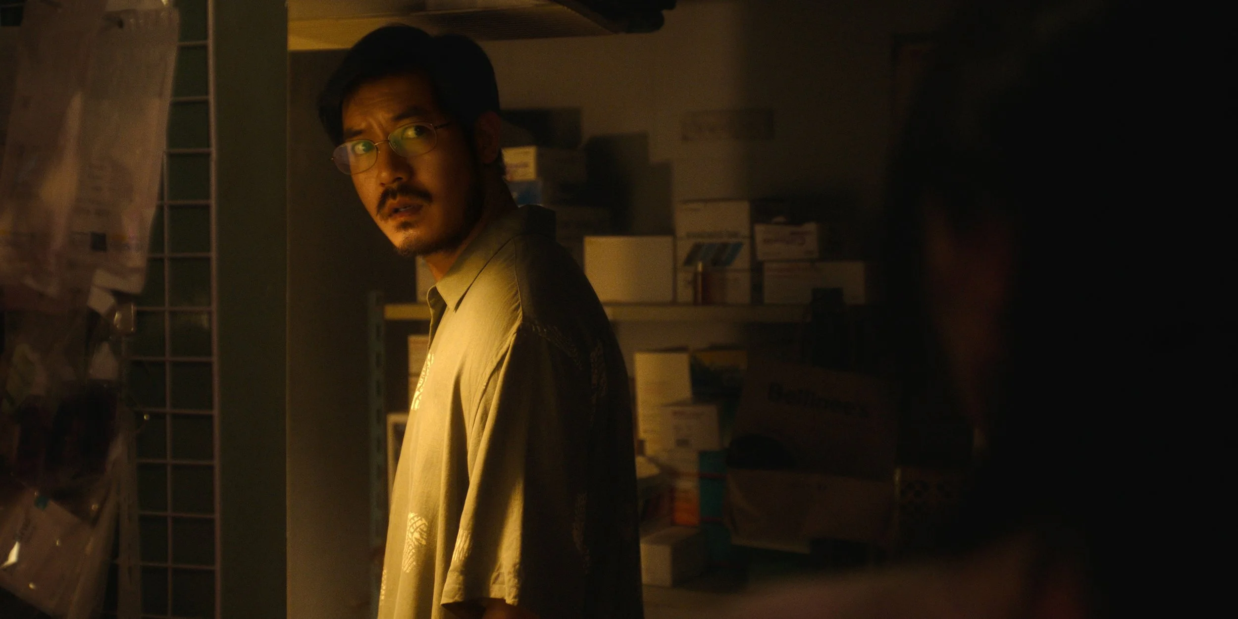 A man with glasses and facial hair looking over his shoulder in a dimly lit storage room or pharmacy with shelves of boxes behind him.