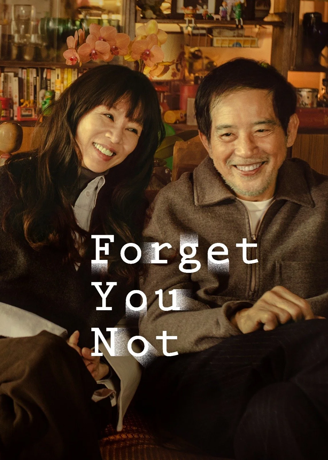 A woman and a man sitting close together, smiling, in a cozy indoor setting with shelves and decorations in the background, with white text overlay that says 'Forget You Not'.