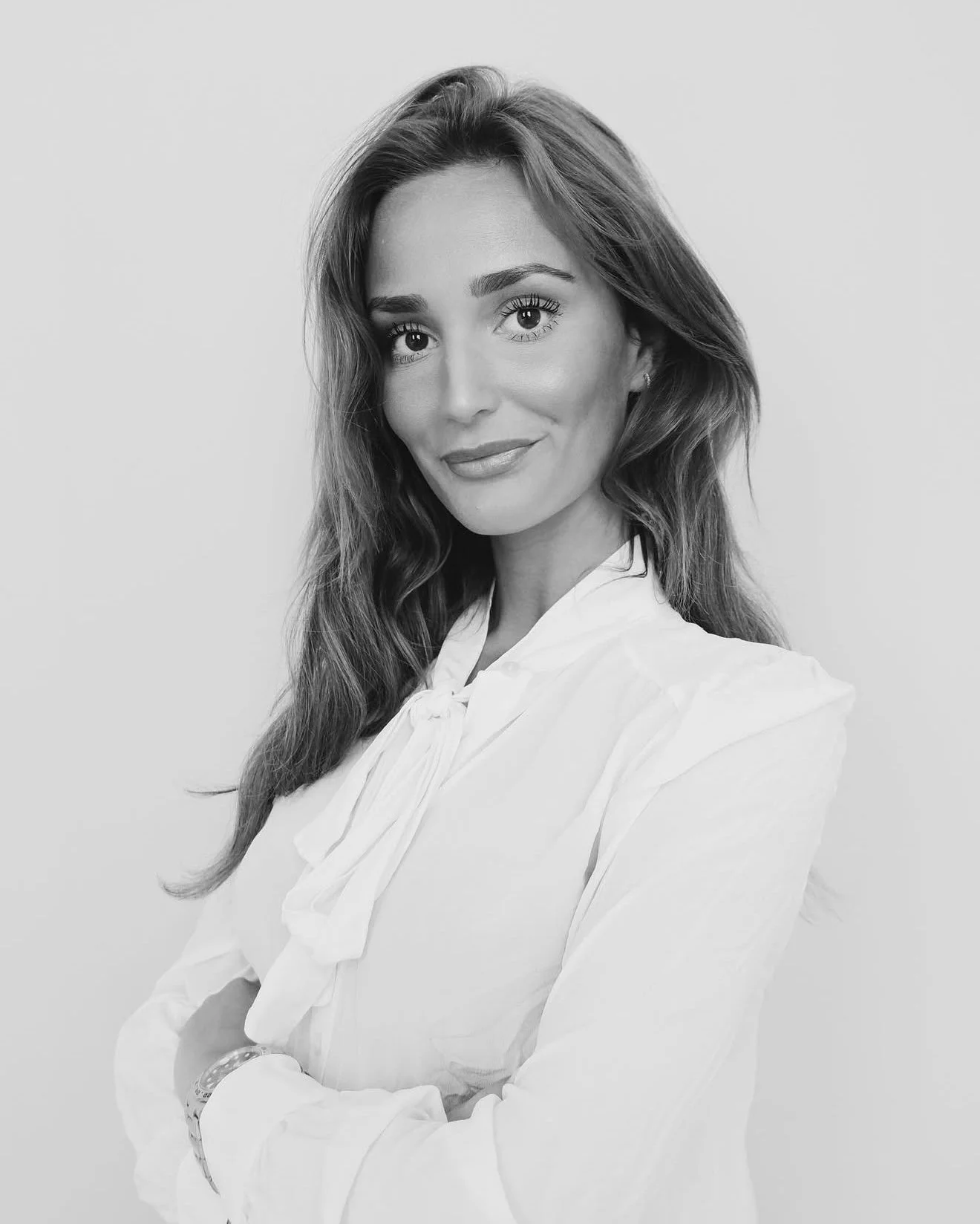 Sort-hvid portrætfoto af en ung kvinde med langt, bølget hår, iført en hvid skjorte, der smiler let mod kameraet med krydsede arme.