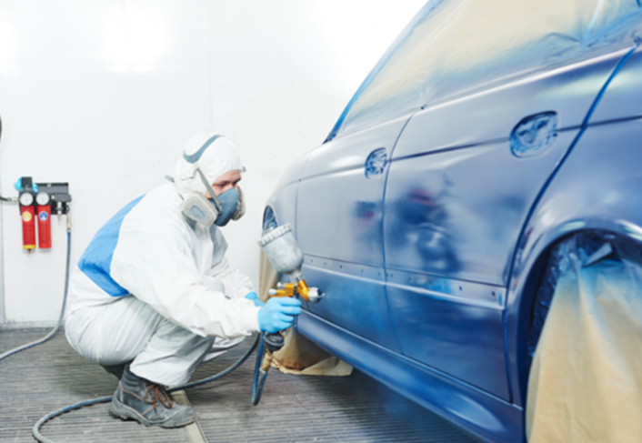 A worker in protective gear spray-paints a blue car in a workshop.