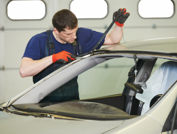 A man wearing a blue shirt and orange gloves working on a car's windshield inside a garage.